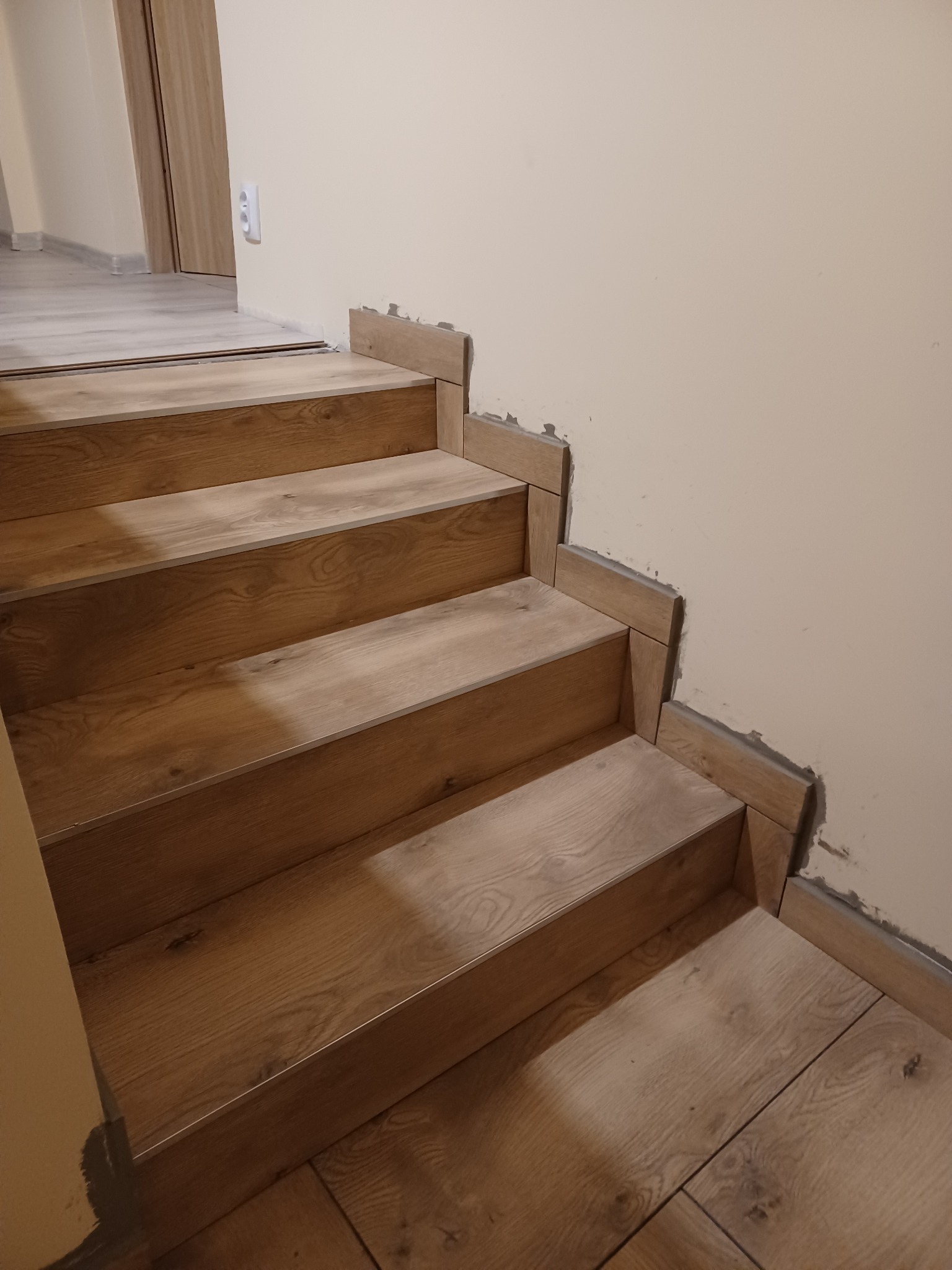 Schody wyłożone panelami imitującymi drewno, z widocznymi śladami kleju i niedokończonymi krawędziami przy ścianie, ukazujące etap prac wykończeniowych.