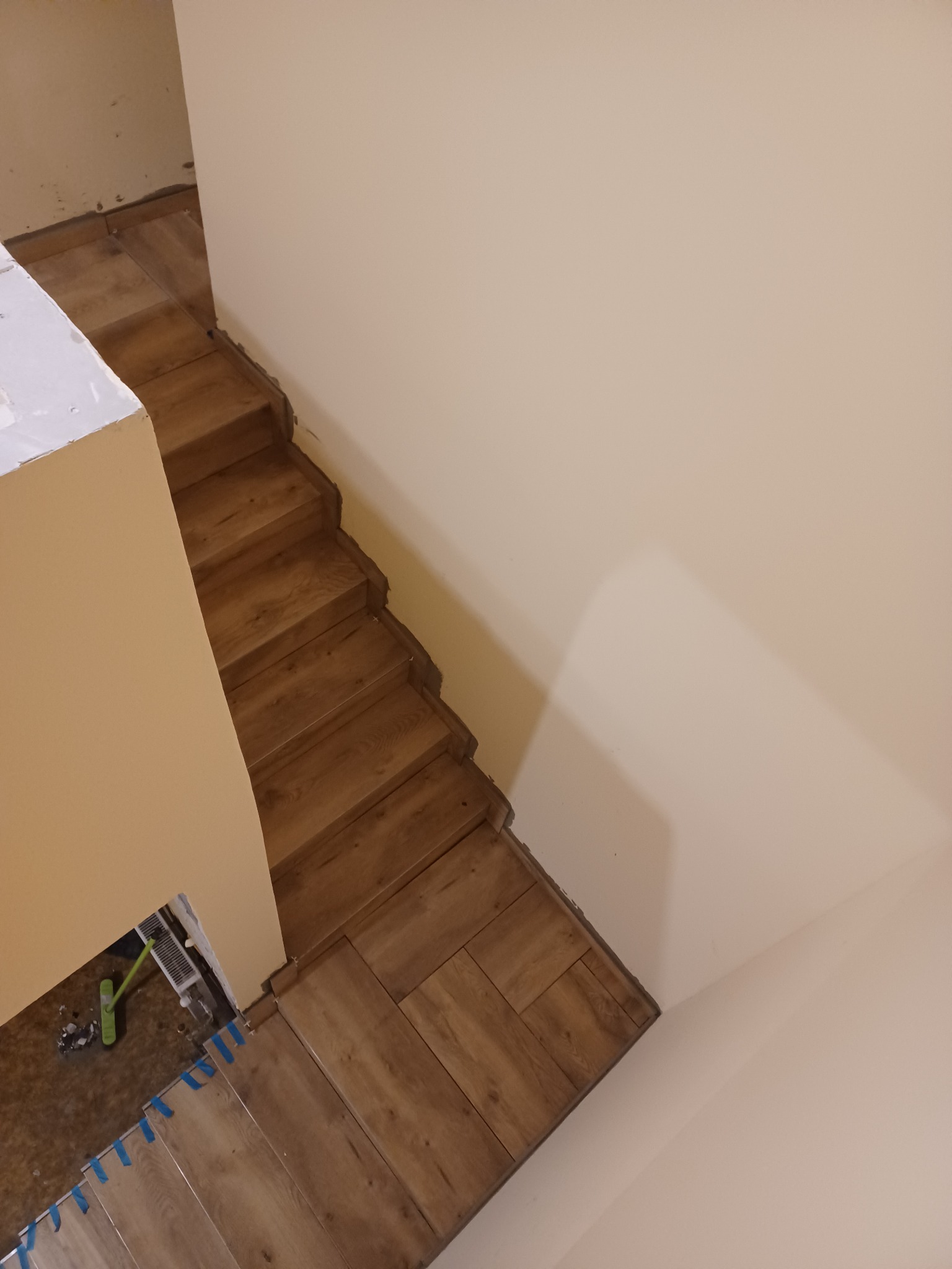 Montaż drewnopodobnych paneli na schodach w trakcie prac wykończeniowych, widoczne nieukończone obrzeża i taśma malarska zabezpieczająca krawędzie, z góry.