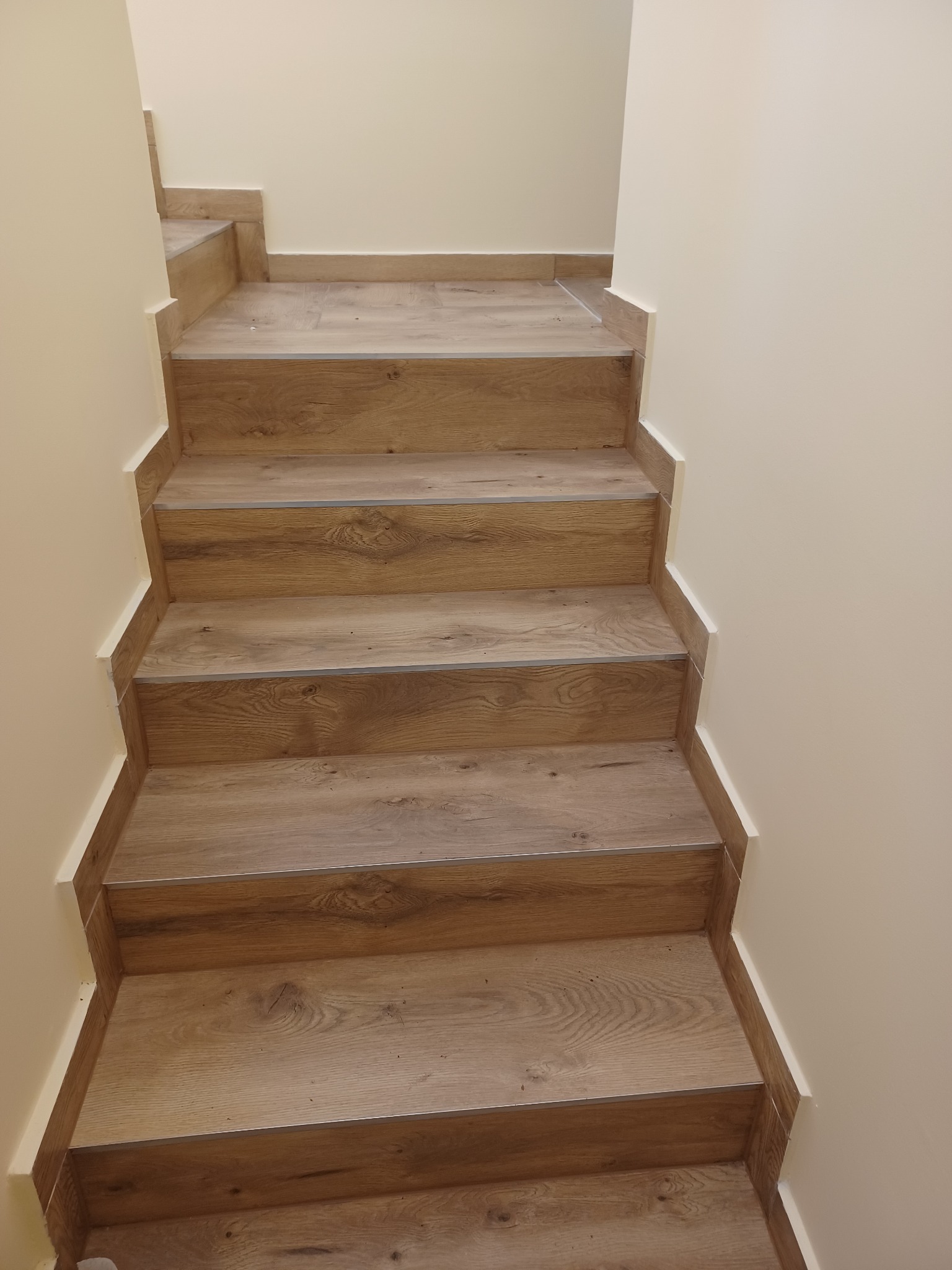 Schody wyłożone panelami imitującymi drewno, z listwami przypodłogowymi w kolorze śmietankowym, prowadzące w górę. Widoczne połączenie paneli na stopniach z delikatną fugą.