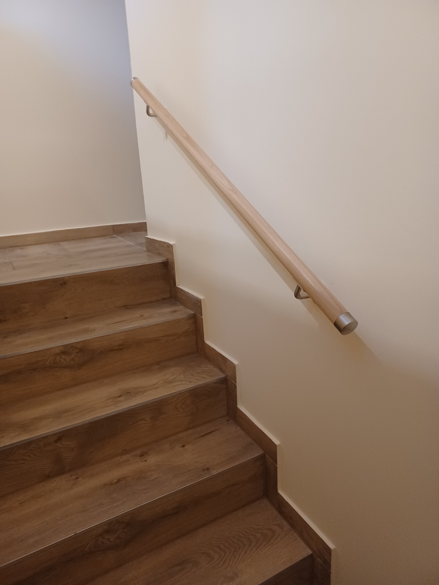 Schody wyłożone panelami imitującymi drewno, z jasną drewnianą poręczą mocowaną do ściany, wykończone listwami przypodłogowymi w kolorze paneli.