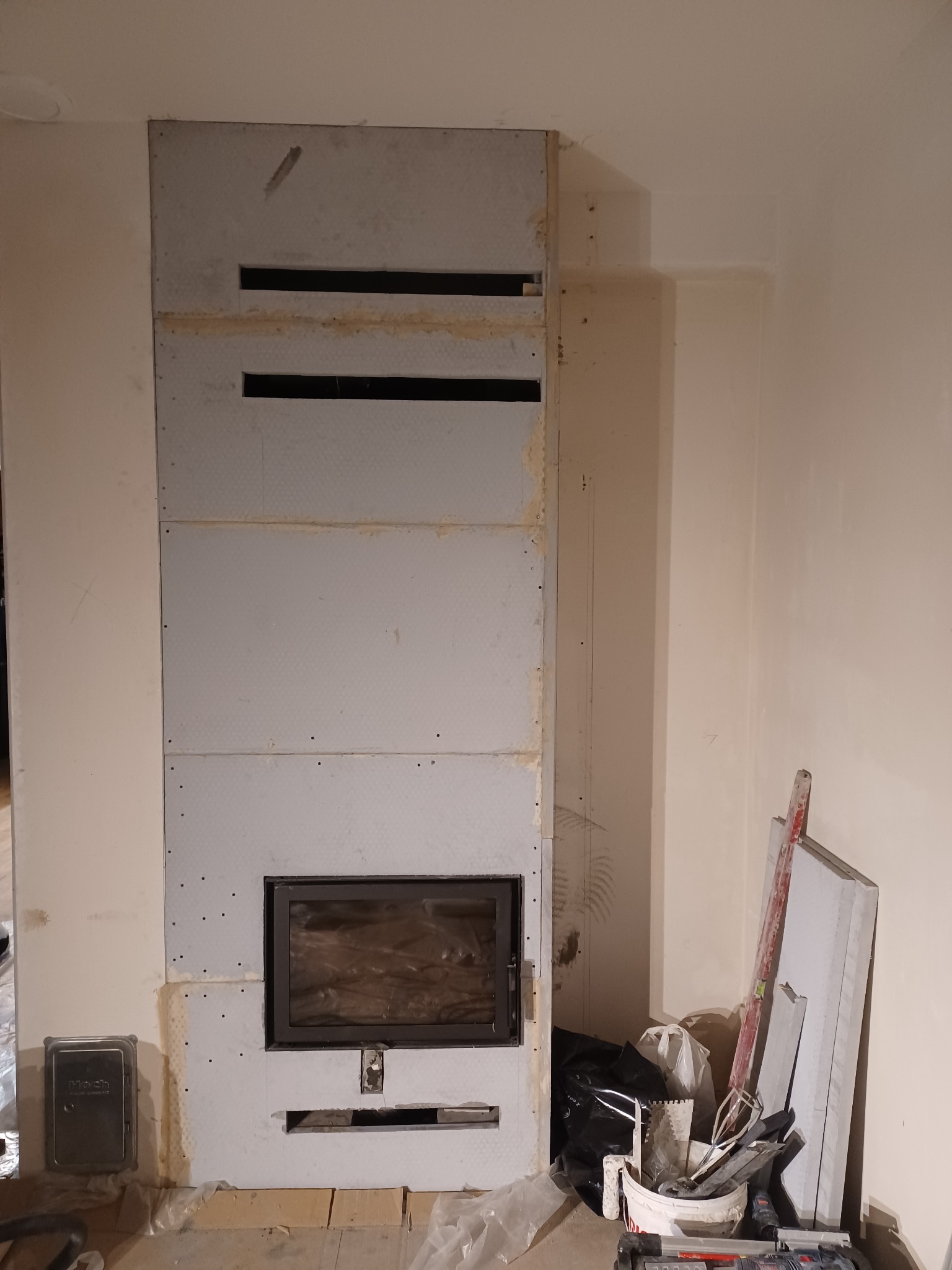 Montaż obudowy kominka z płyt gipsowo-kartonowych, widoczne otwory wentylacyjne i wkład kominkowy, narzędzia i materiały budowlane w pobliżu.