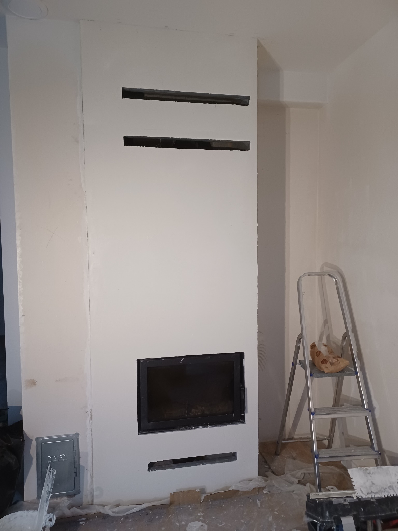 Wnętrze w trakcie wykańczania z kominkiem obudowanym płytami gipsowo-kartonowymi ze szpachlowanymi łączeniami, widoczne trzy poziome otwory wentylacyjne nad paleniskiem oraz drabina aluminiowa...