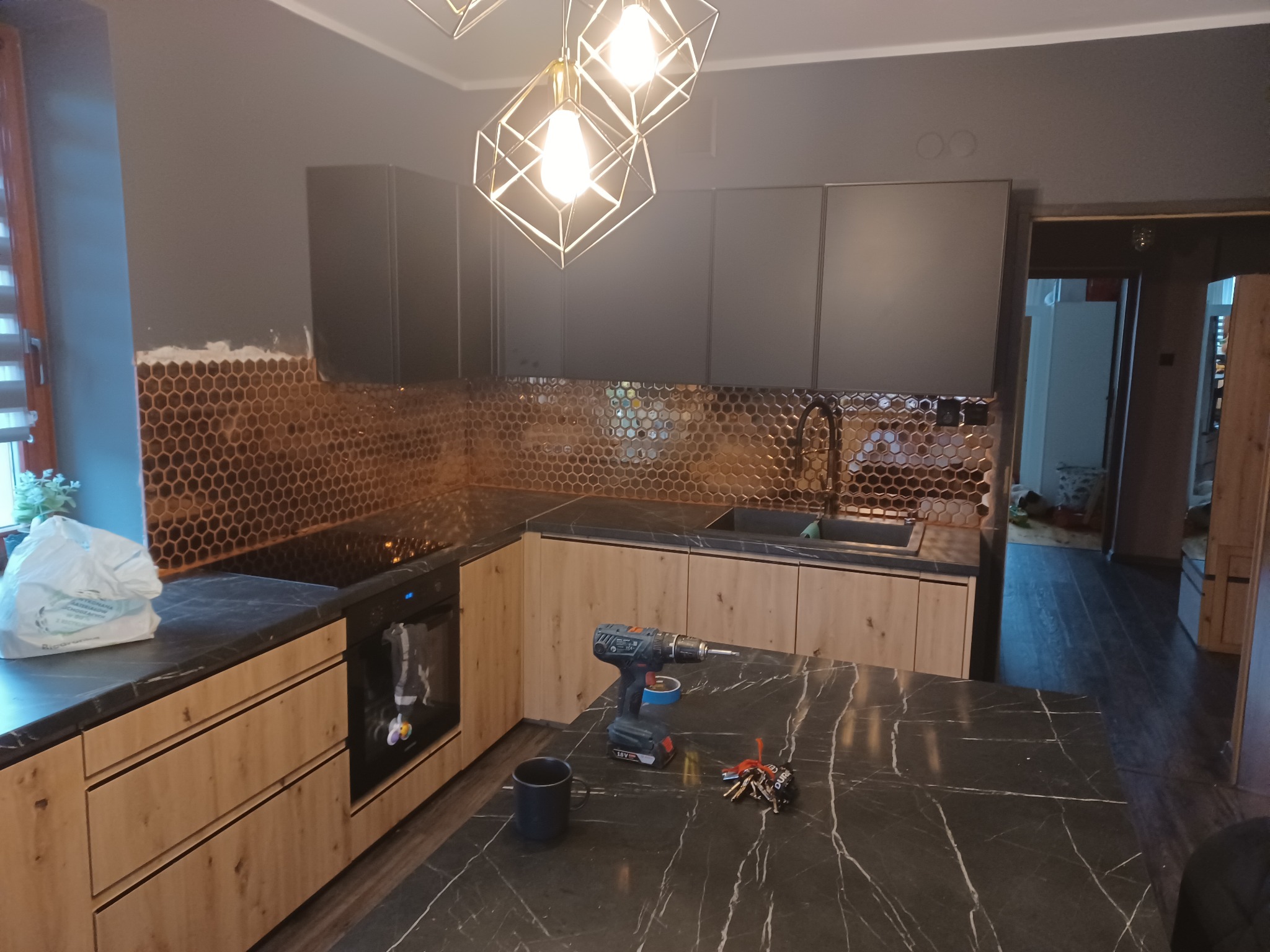 Nowoczesna kuchnia w trakcie prac wykończeniowych: ciemne szafki górne, drewnopodobne dolne, mozaika w kształcie plastra miodu na ścianie, na blacie wiertarka akumulatorowa, kubek i klucze.