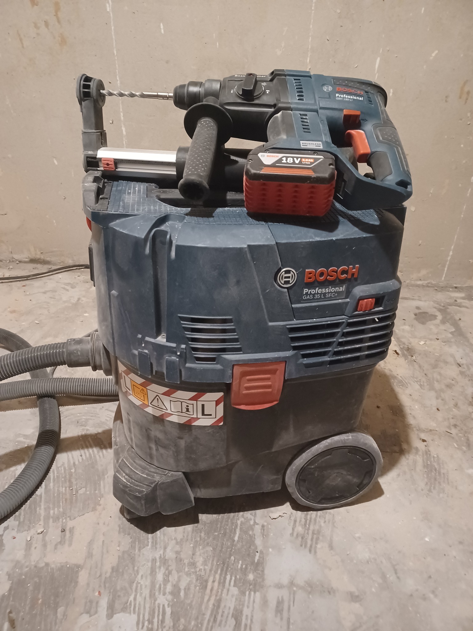 Profesjonalny odkurzacz budowlany Bosch GAS 35 L SFC+ z podłączonym młotem udarowym Bosch GBH 180-LI z wiertłem, stojący na betonowej podłodze w trakcie lub po pracy.