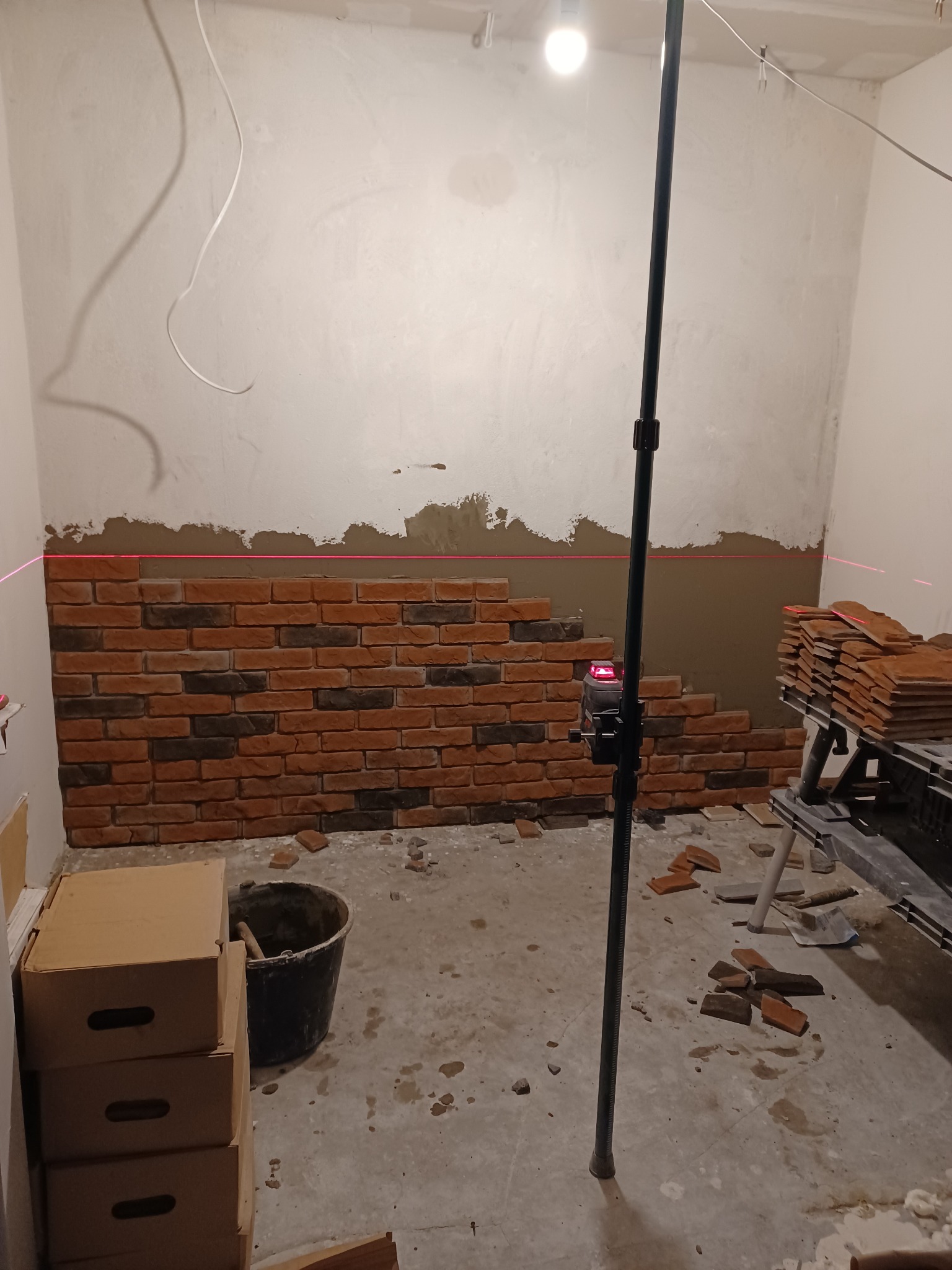 Fragment ściany z częściowo ułożoną dekoracyjną cegłą, poziom wyznaczony laserem, widoczne narzędzia i materiały budowlane w pomieszczeniu w trakcie prac.