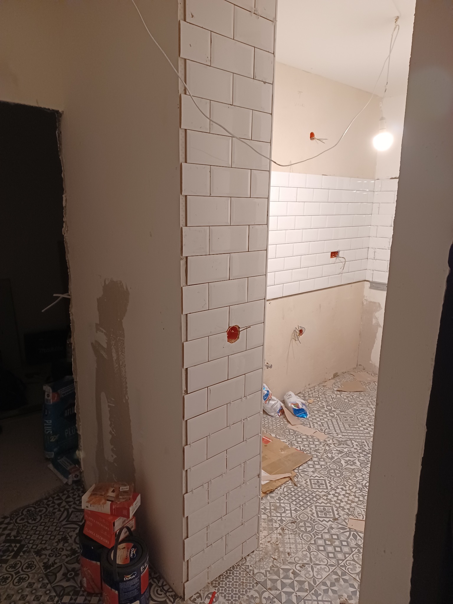 Wnętrze łazienki w trakcie prac: białe, prostokątne płytki ułożone do połowy ściany, widoczne puszki elektryczne, podłoga z wzorzystych płytek, worki z zaprawą i farby.