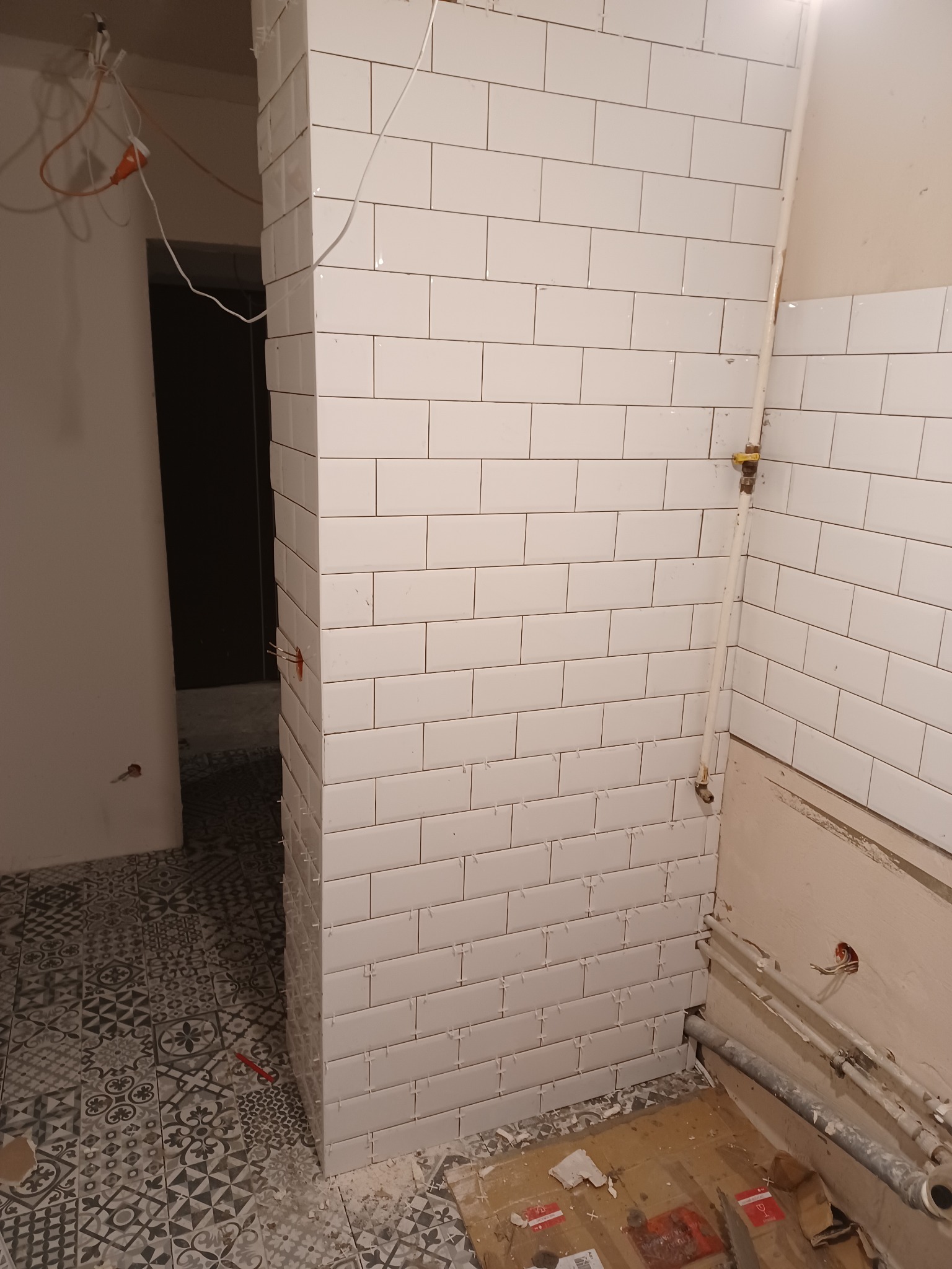 Białe, prostokątne płytki ułożone na ścianie w łazience w trakcie remontu, widoczne krzyżyki dystansowe, rury i przewody elektryczne, wzorzysta podłoga.