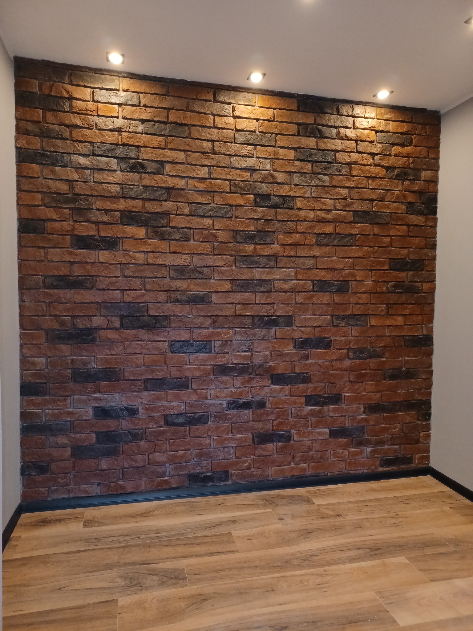 Ściana z dekoracyjnej cegły w odcieniach brązu i czerni, oświetlona punktowymi lampami sufitowymi, z widoczną podłogą z paneli imitujących drewno.