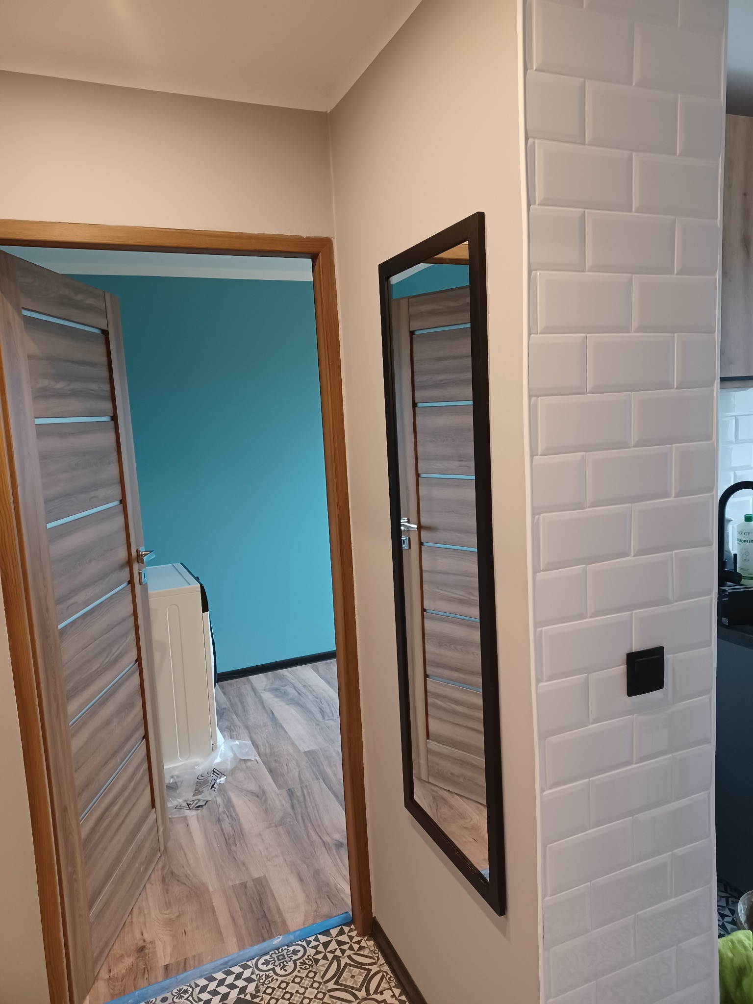 Fragment korytarza po remoncie: białe płytki typu metro, czarne lustro w ramie, widok na łazienkę z turkusową ścianą i drewnopodobną podłogą, wzorzysta podłoga na granicy pomieszczeń.