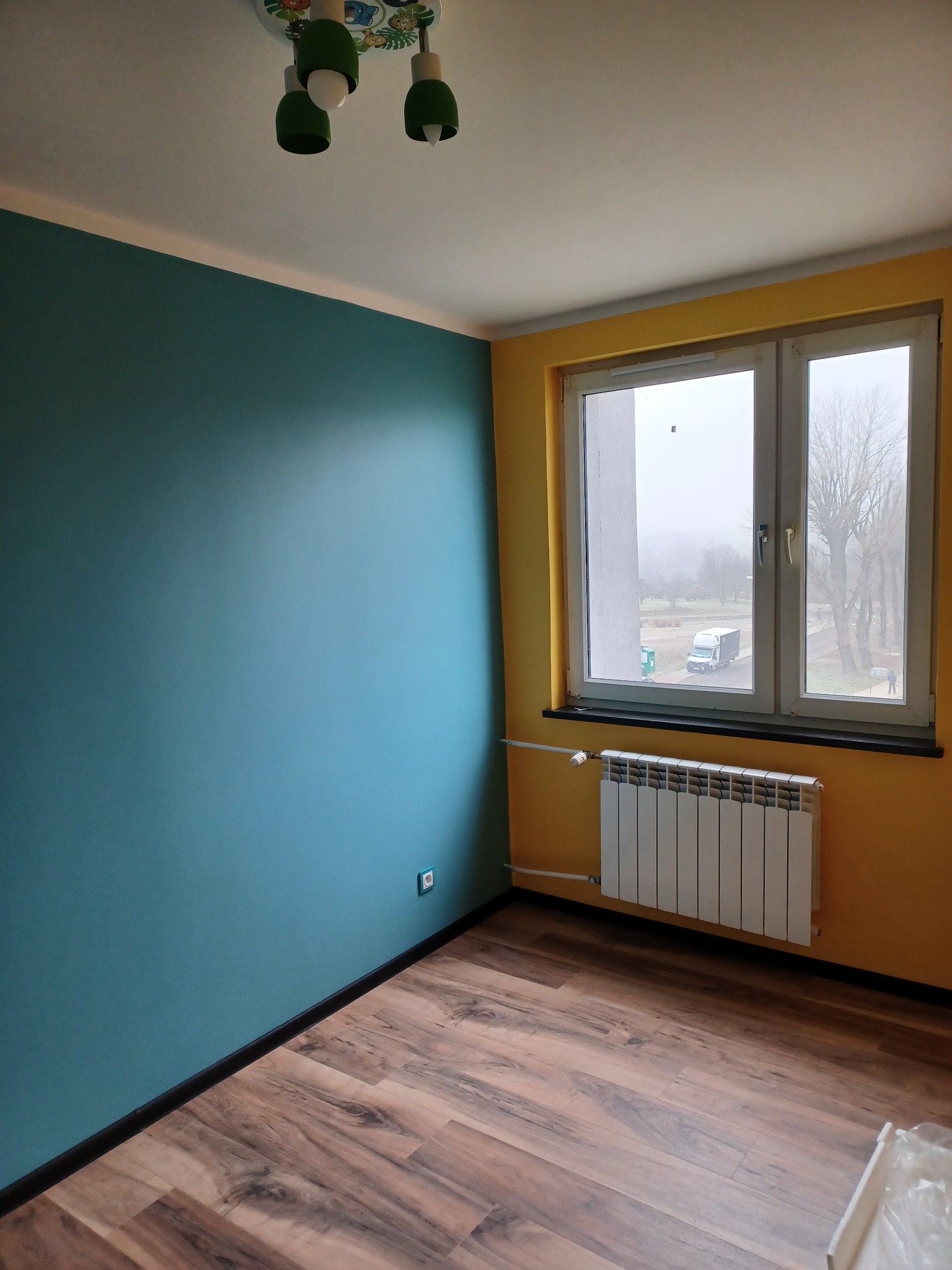 Pomalowane na turkusowo i żółto wnętrze pokoju z panelami podłogowymi, białym grzejnikiem i oknem z widokiem na pochmurny krajobraz miejski.