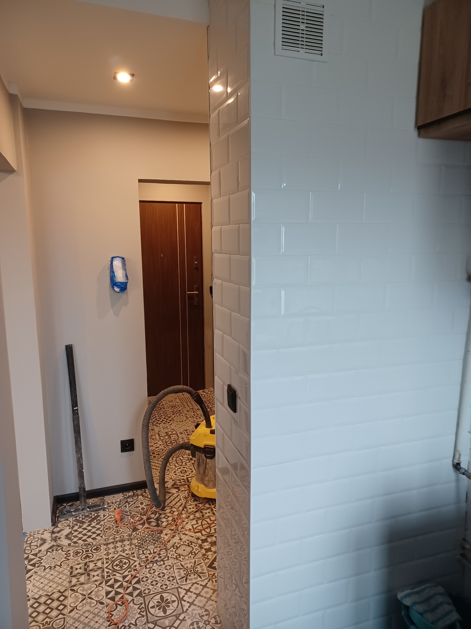 Białe, błyszczące płytki ułożone na ścianie w kształcie cegiełek, widoczny fragment podłogi z wzorzystymi płytkami, odkurzacz przemysłowy, oraz fragment korytarza z brązowymi drzwiami i niebieskim...