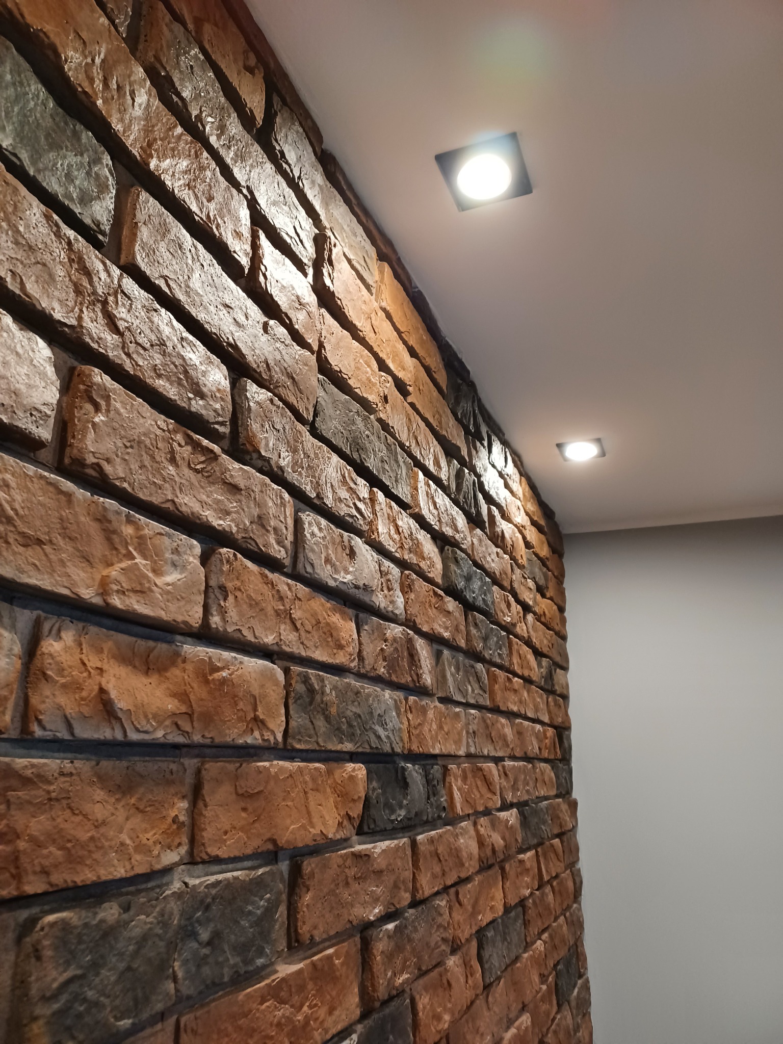 Dekoracyjna ściana z cegły klinkierowej w odcieniach brązu i szarości, oświetlona dwoma punktowymi lampami sufitowymi, widok z dołu pod kątem.