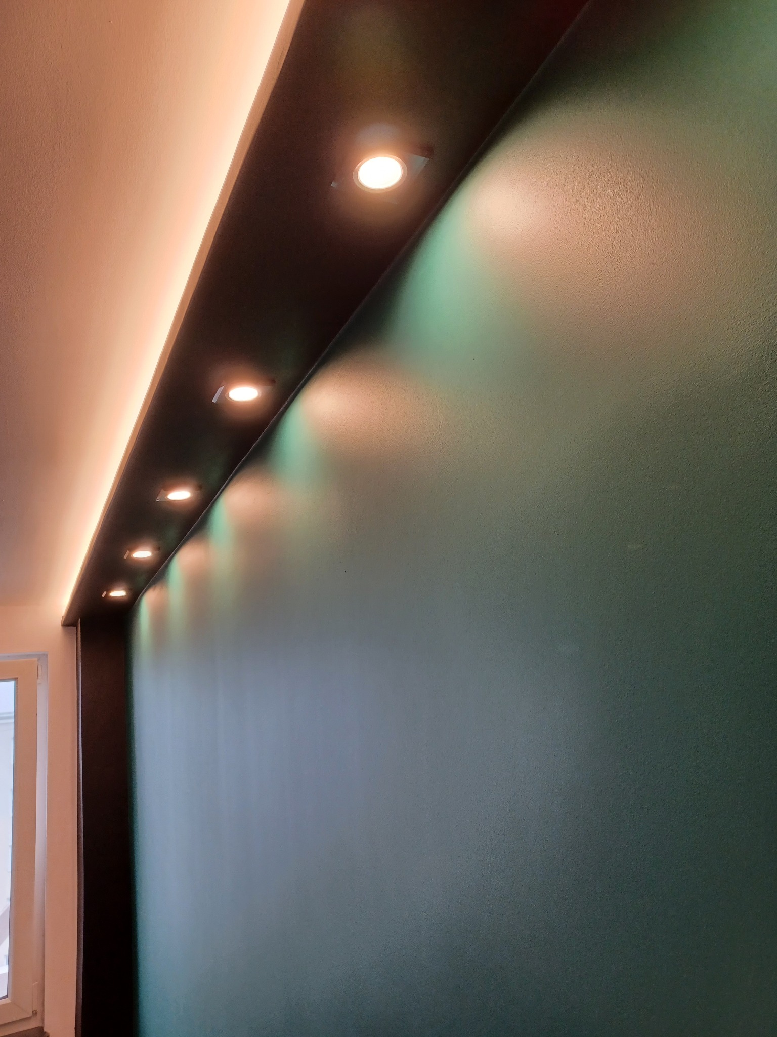 Ściana w odcieniach zieleni z rzędem wbudowanych, punktowych lamp LED w czarnej listwie, oświetlających sufit i ścianę.