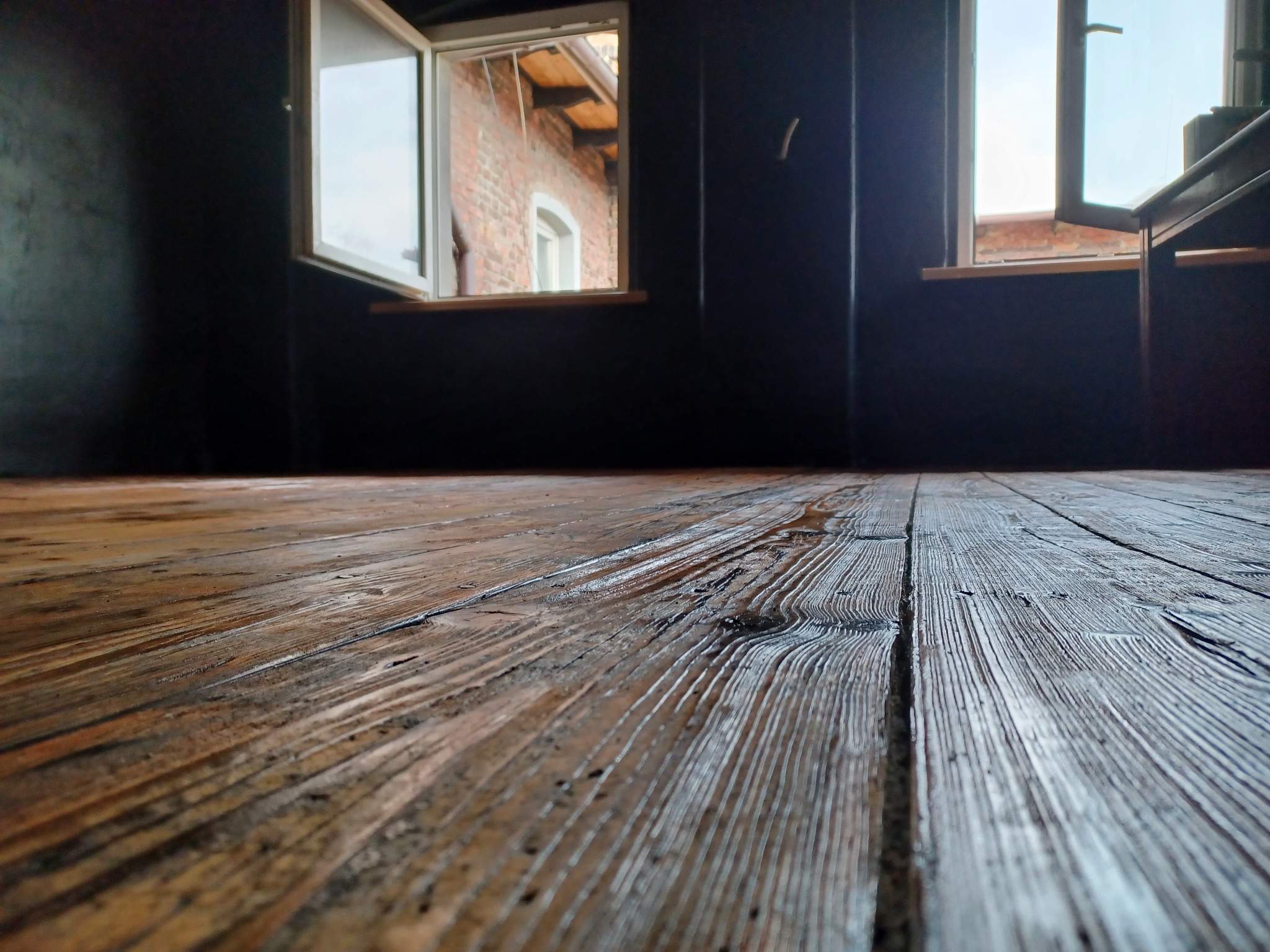 Odrestaurowana drewniana podłoga z widocznymi słojami i śladami po lakierowaniu, na tle ciemnych ścian i otwartych okien z widokiem na ceglany budynek.