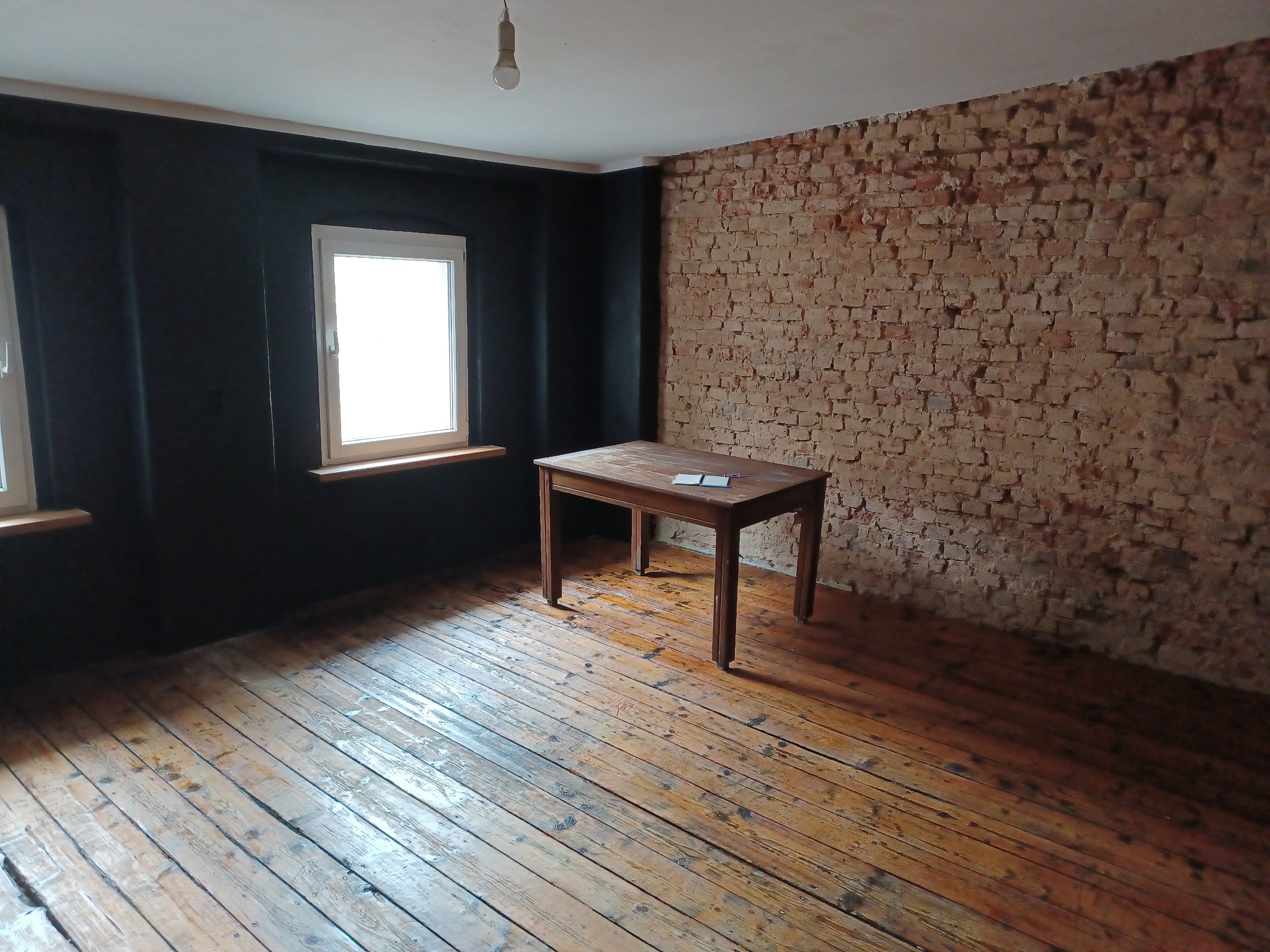 Pokój w trakcie remontu: ściana z surowej cegły kontrastuje z pomalowaną na czarno ścianą, drewniana podłoga z widocznymi śladami użytkowania i stary stół na środku pomieszczenia.