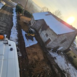 Auto-komplex - Żółty mini-koparka Volvo na białej macie, wykopany rów wzdłuż budynku z szarego betonu, częściowo pokrytego śniegiem, w słoneczny dzień.