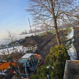 Auto-komplex - Pomarańczowa koparka kołowa w trakcie prac ziemnych na zasnieżonym terenie, widok z góry na wykop i operatora w kabinie. Zima, błękitne niebo.