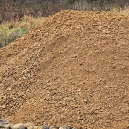 Auto-komplex - Góra piasku i kamieni na tle zarośli. Widoczne nierówności terenu i mieszanka materiałów. Zastosowanie w robotach ziemnych.