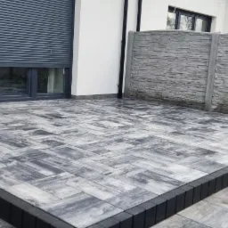 Nowoczesny taras z szaro-białej kostki ułożonej we wzór jodełki, z czarnymi obrzeżami schodów, otoczony betonowym ogrodzeniem imitującym kamień.