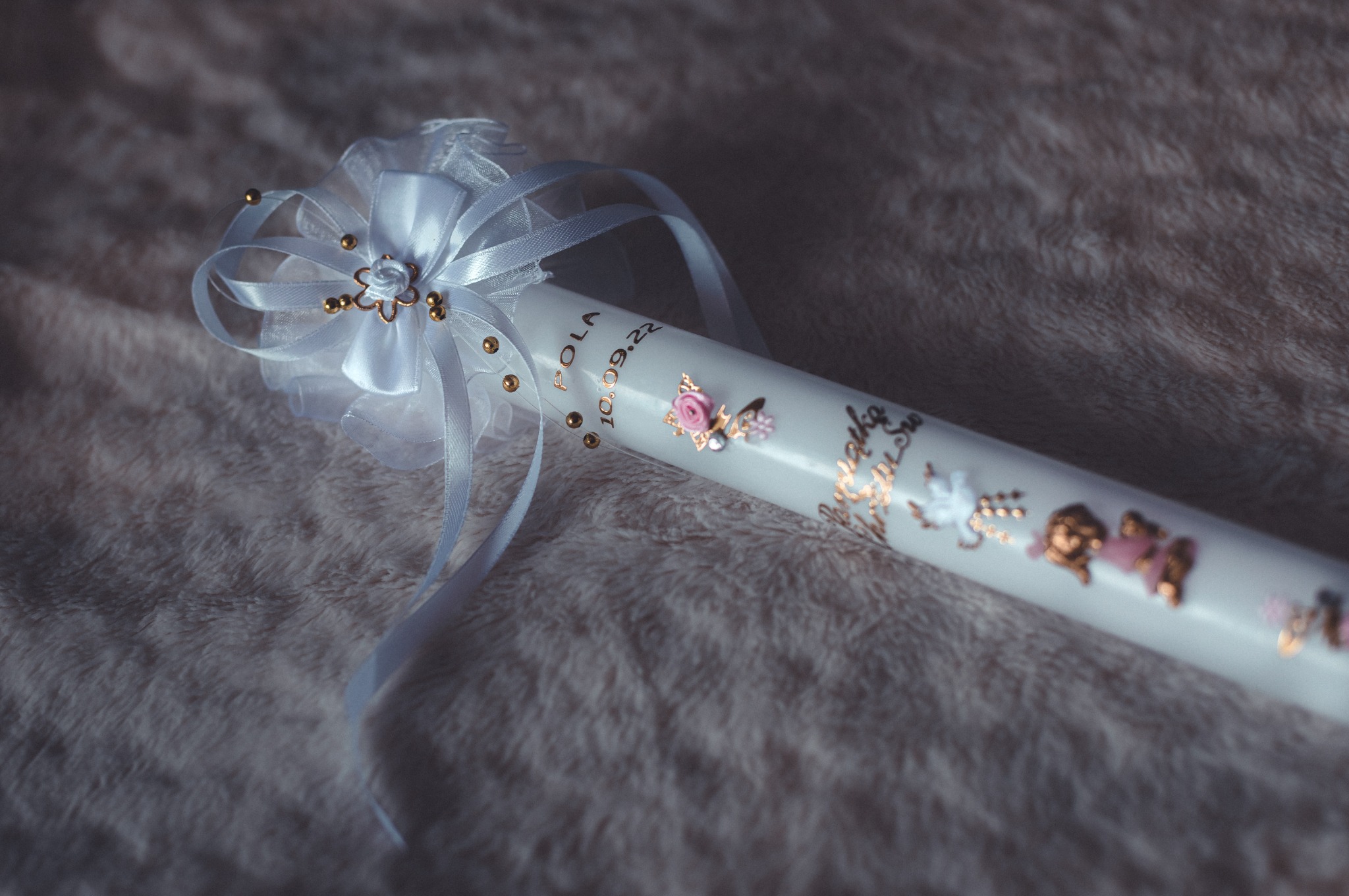 Ozdobna, biała świeca z imieniem 'Pola' i datą '10.09.22', udekorowana złotymi elementami i białymi wstążkami, leżąca na miękkim, puszystym tle.