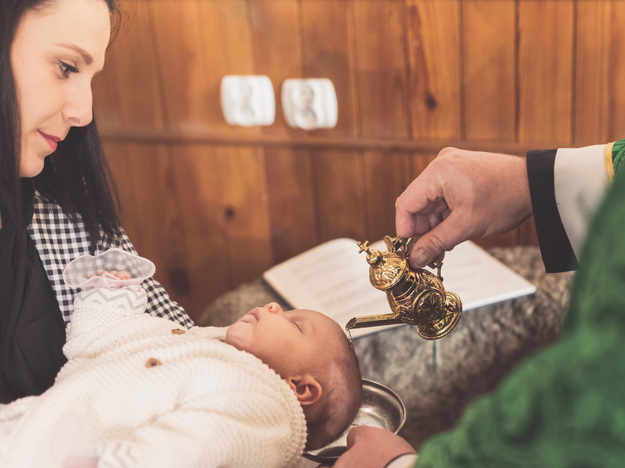 Ksiądz polewa wodą z ozdobnej złotej karafki głowę śpiącego niemowlęcia podczas ceremonii chrztu, matka dziecka przygląda się z troską.