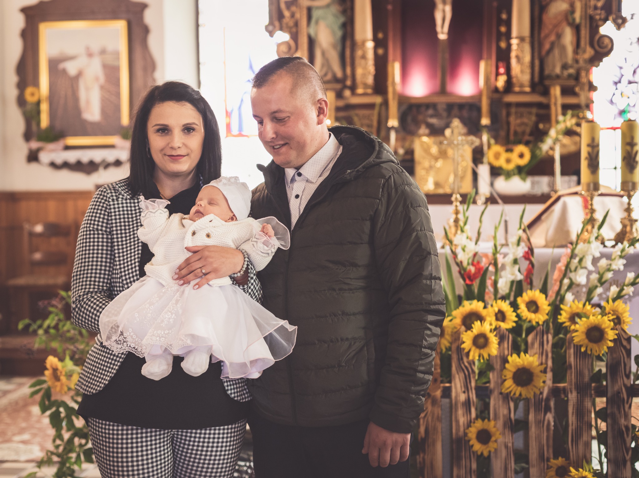 Rodzice z dzieckiem w stroju do chrztu w kościele, dekoracje z kwiatów słonecznika, portret rodzinny.