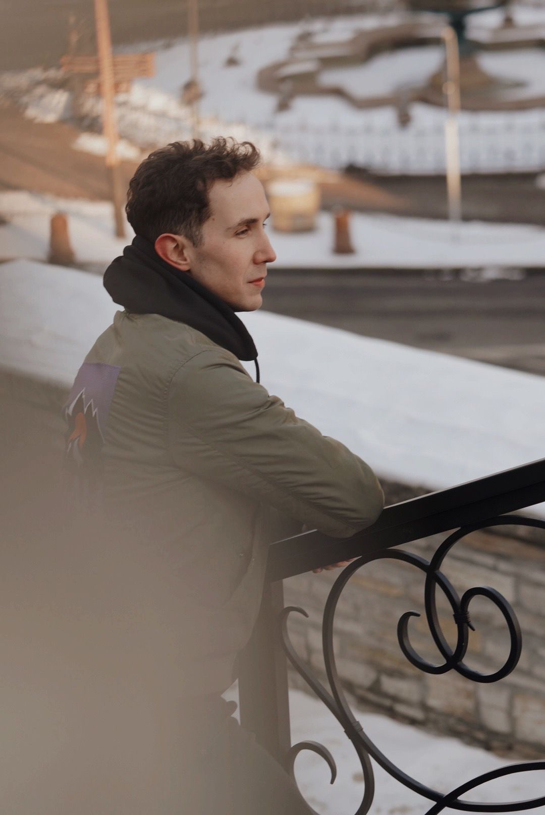 Młody mężczyzna w profilu, oparty o ozdobną, metalową balustradę na tle zimowego krajobrazu miejskiego, ubrany w zieloną kurtkę z naszywką górską.