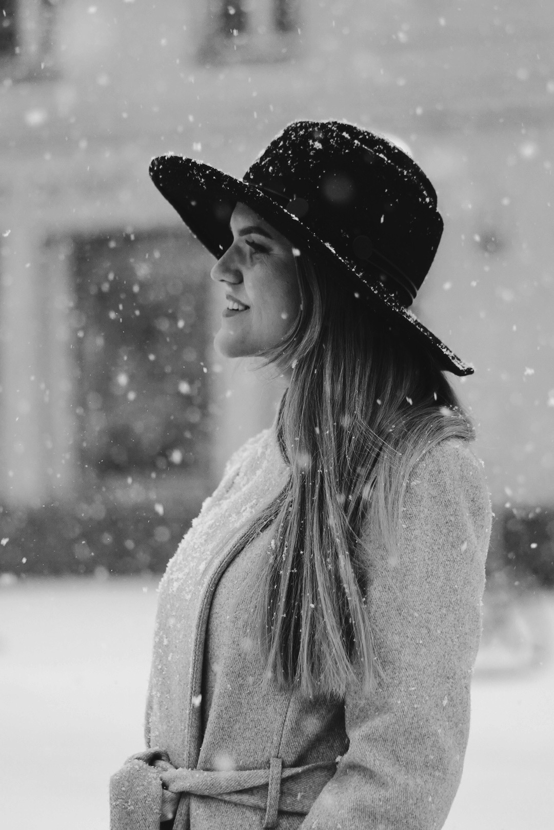 Portret kobiety w kapeluszu, uchwycony w monochromatycznej tonacji podczas padającego śniegu, z delikatnym uśmiechem i profilowym ujęciem.