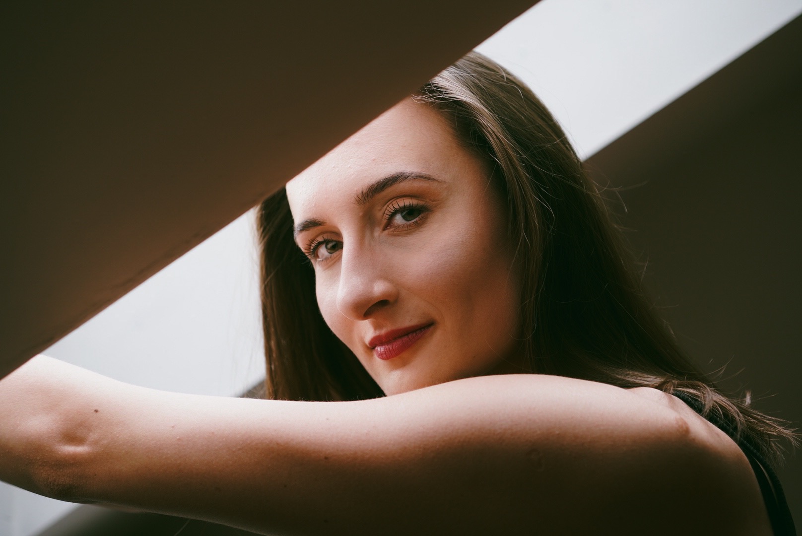 Portret uśmiechniętej kobiety z długimi brązowymi włosami, oświetlonej przez naturalne światło wpadające przez okno, z nietypowym kadrowaniem.