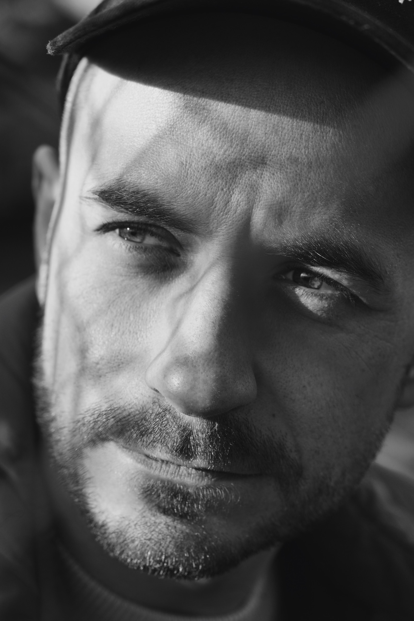 Czarno-biały portret mężczyzny z brodą i czapką, oświetlony z góry, tworzący cień na twarzy.