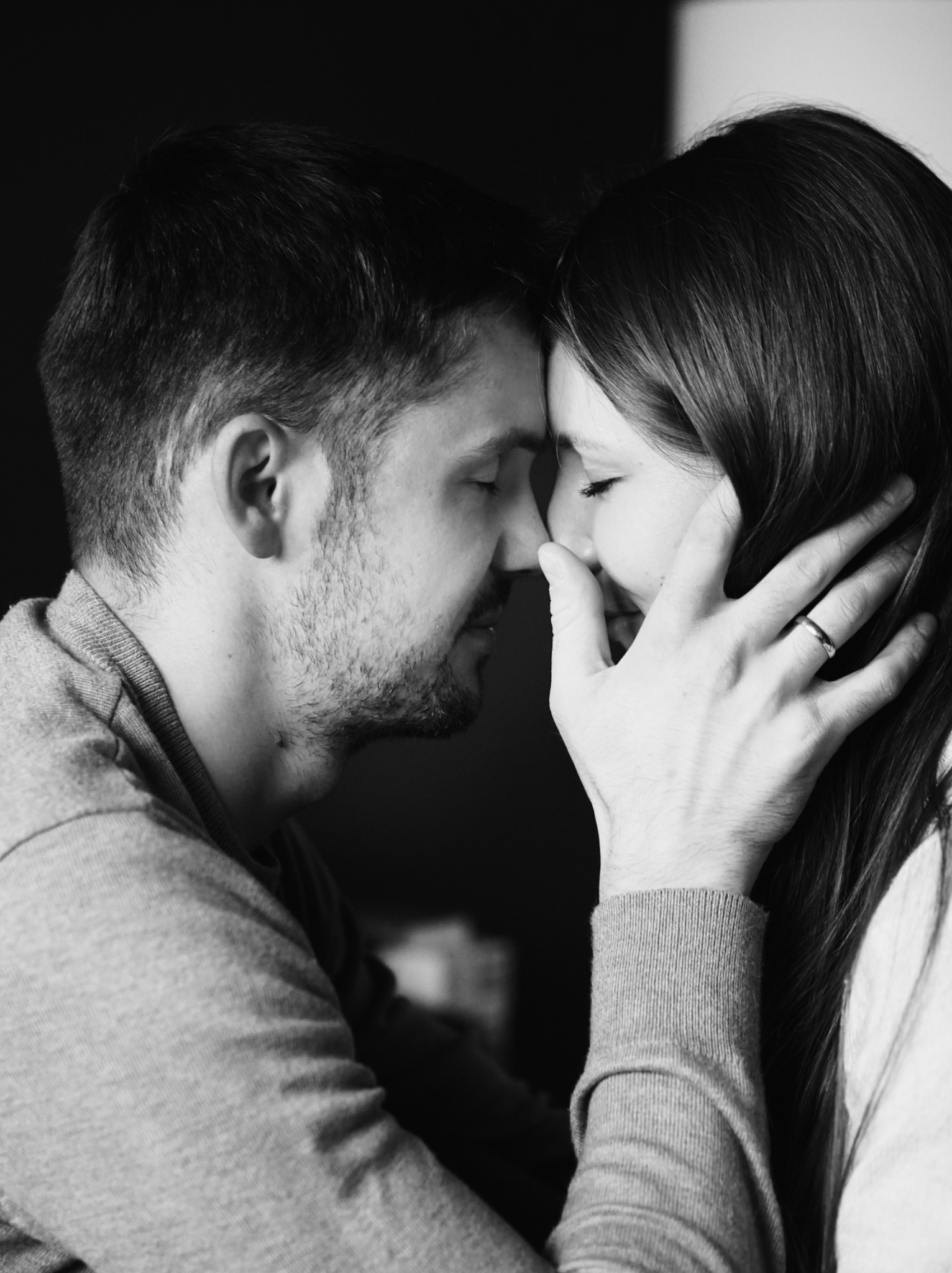 Czarno-białe zbliżenie twarzy pary, dotykających się nosami, z dłonią mężczyzny z obrączką delikatnie obejmującą policzek kobiety, zamknięte oczy, subtelne światło.