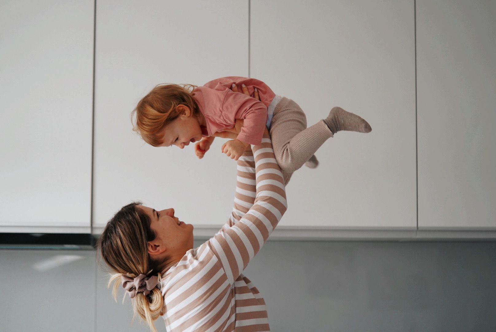 Uśmiechnięta mama w bluzce w paski podnosi wysoko w górę roześmianą, rudowłosą córeczkę w różowej bluzce i beżowych legginsach na tle białych szafek.