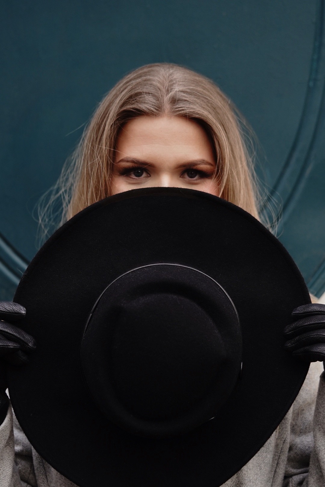 Portret kobiety z blond włosami i wyrazistym makijażem oczu, częściowo zasłoniętej dużym, czarnym kapeluszem i skórzanymi rękawiczkami na tle turkusowych drzwi.