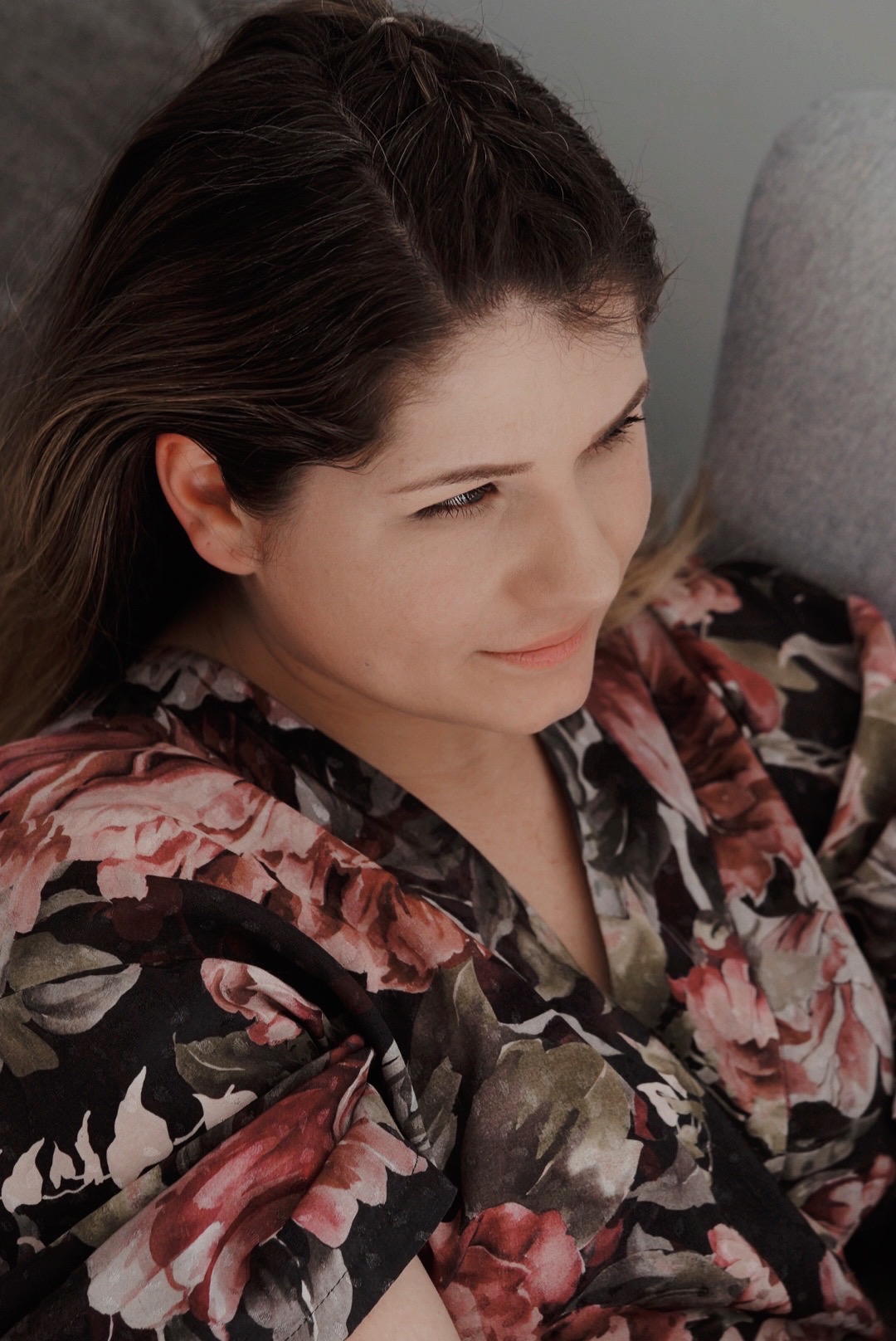 Portret kobiety z profilu, ubranej w kwiecisty szlafrok, z delikatnym uśmiechem i spojrzeniem skierowanym w dół.