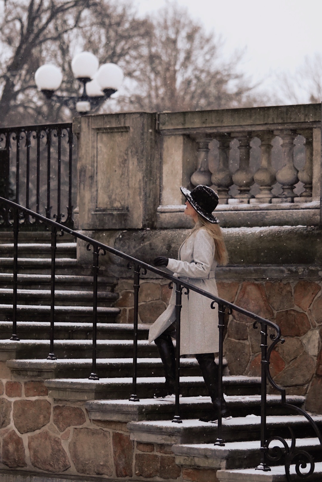 Elegancka kobieta w płaszczu i kapeluszu wchodzi po kamiennych schodach w zimowej scenerii, z delikatnym opadem śniegu i ozdobnymi latarniami w tle.