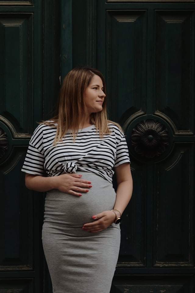 Uśmiechnięta kobieta w ciąży, ubrana w szaro-białą koszulkę w paski i szarą sukienkę, pozuje na tle ciemnozielonych, zdobionych drzwi.