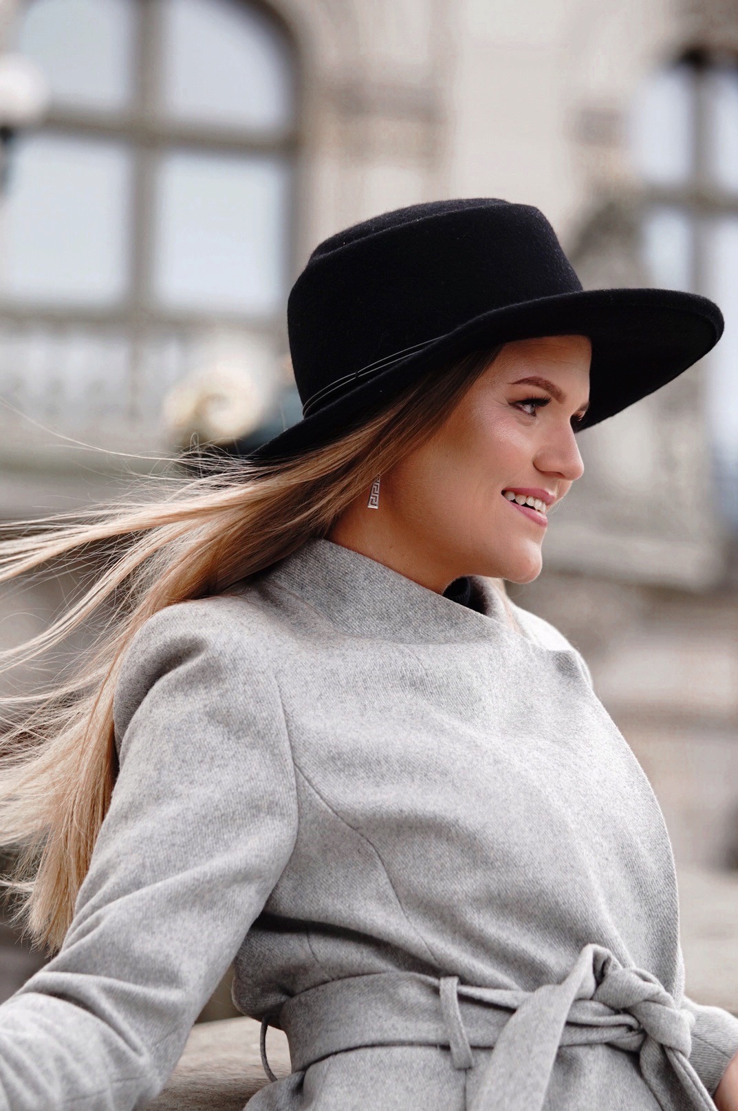 Portret uśmiechniętej kobiety w szarym płaszczu i czarnym kapeluszu, z rozwianymi blond włosami, sfotografowanej w Bielsku-Białej na tle miejskiej architektury.