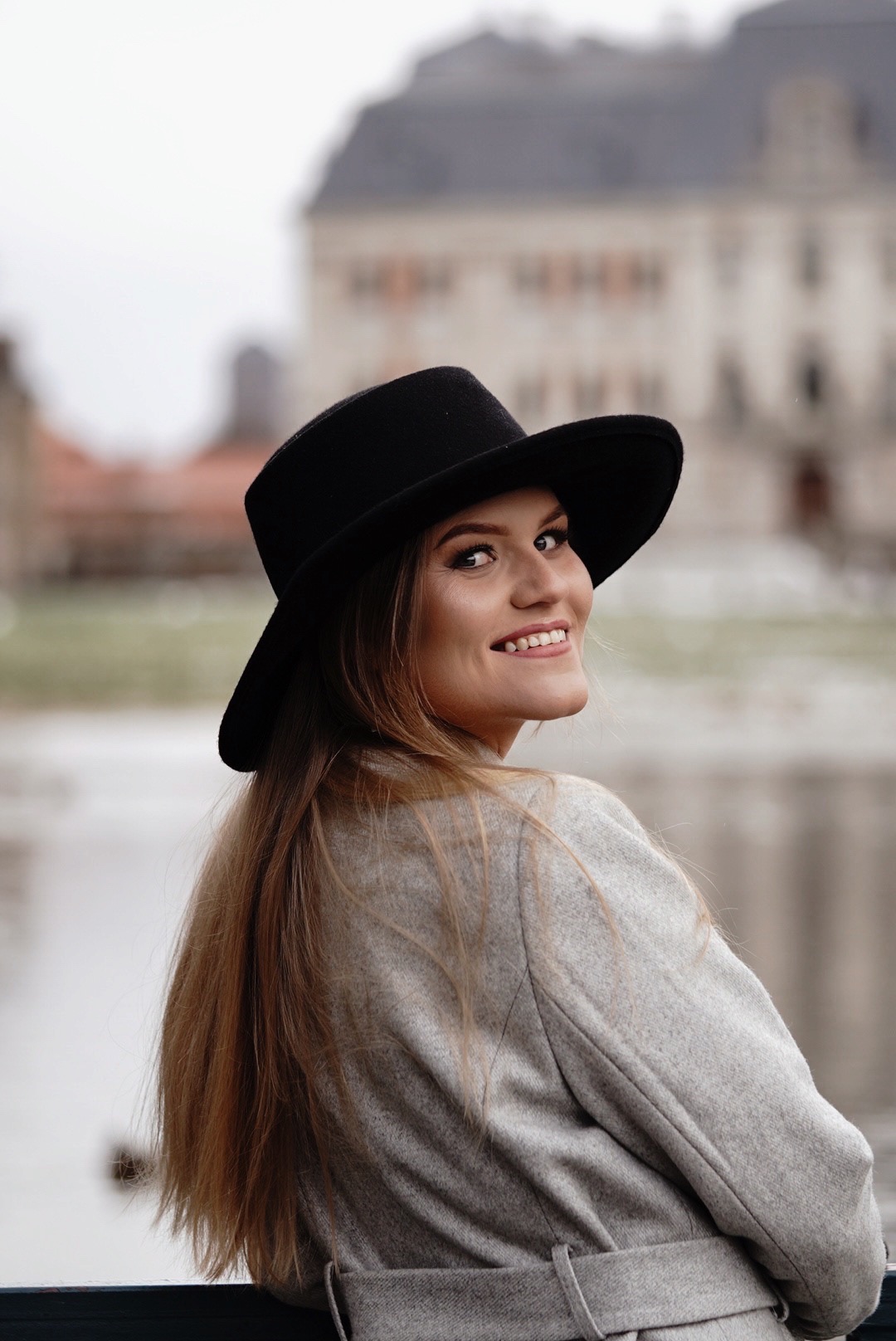 Uśmiechnięta kobieta w czarnym kapeluszu i szarym płaszczu, odwrócona przez ramię, z budynkiem w tle.