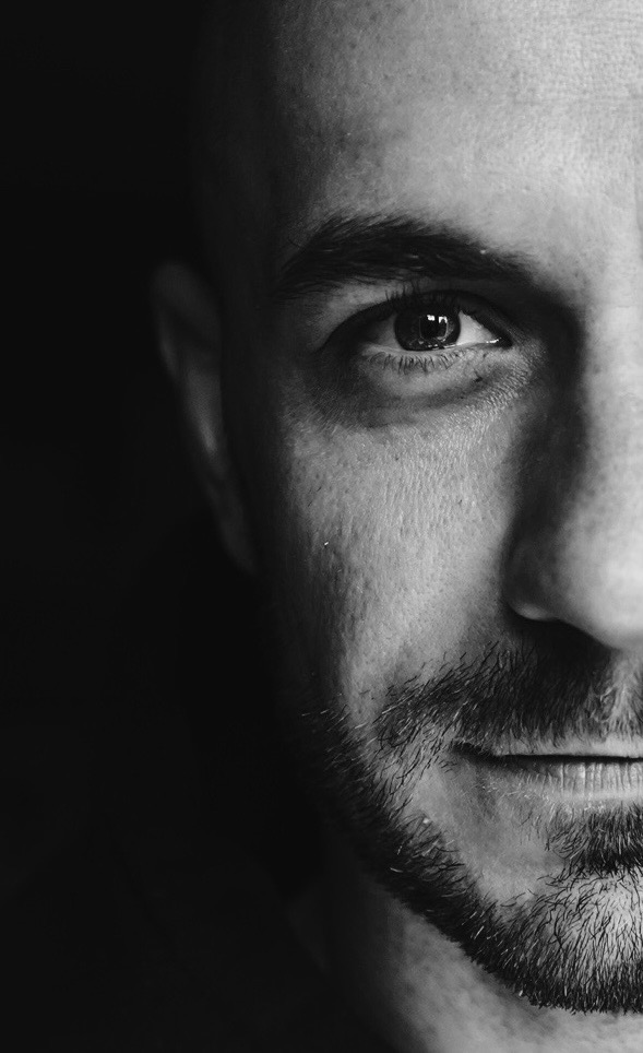 Czarno-biały portret mężczyzny z bliska, widoczna połowa twarzy, z ostrym światłem padającym na oko i brodę, tworząc dramatyczny efekt.