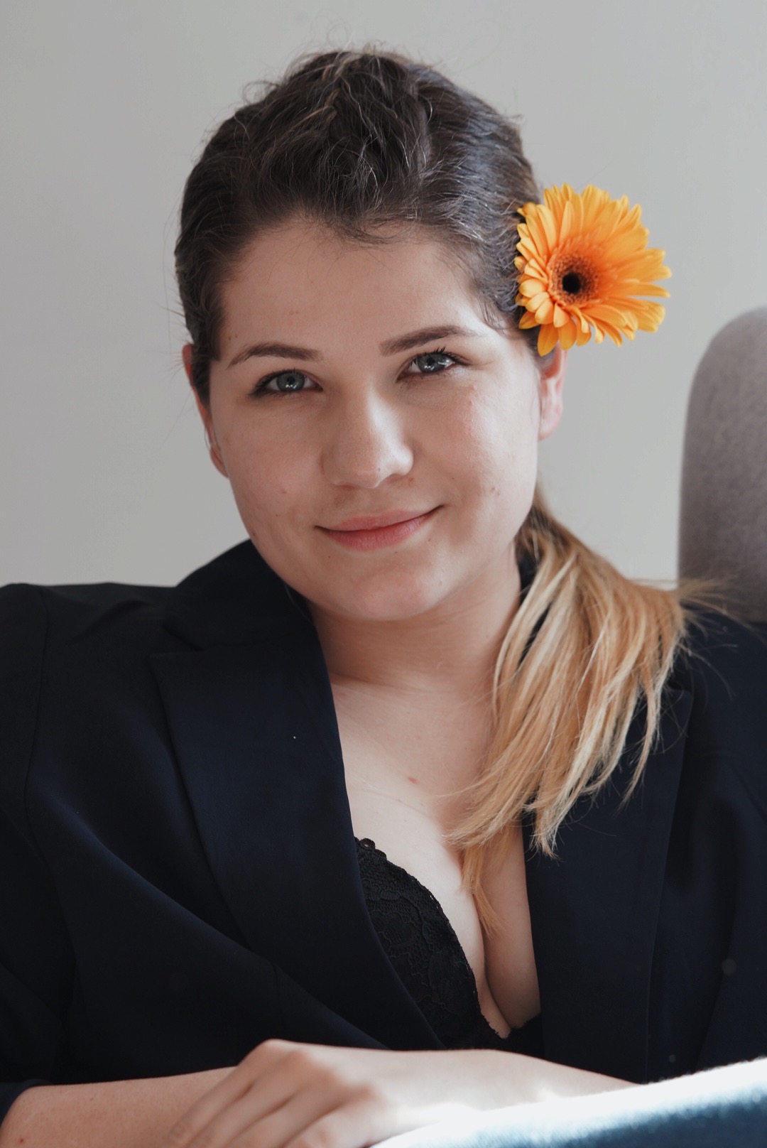 Portret uśmiechniętej kobiety w czarnej marynarce z pomarańczową gerberą we włosach, blond kosmyk włosów.