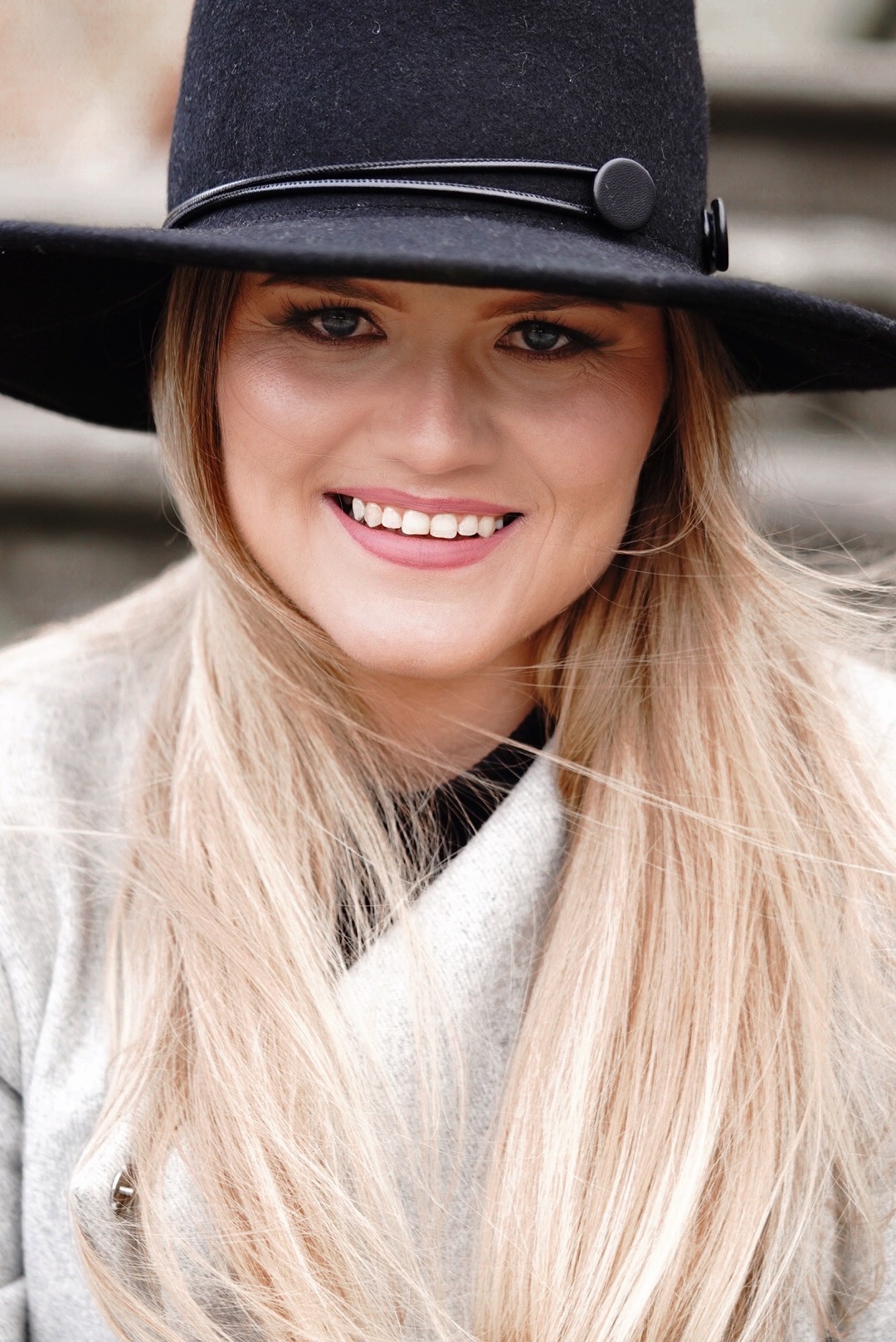 Portret uśmiechniętej kobiety w czarnym kapeluszu z szerokim rondem i jasnym płaszczu, z rozwianymi blond włosami, na rozmytym tle.