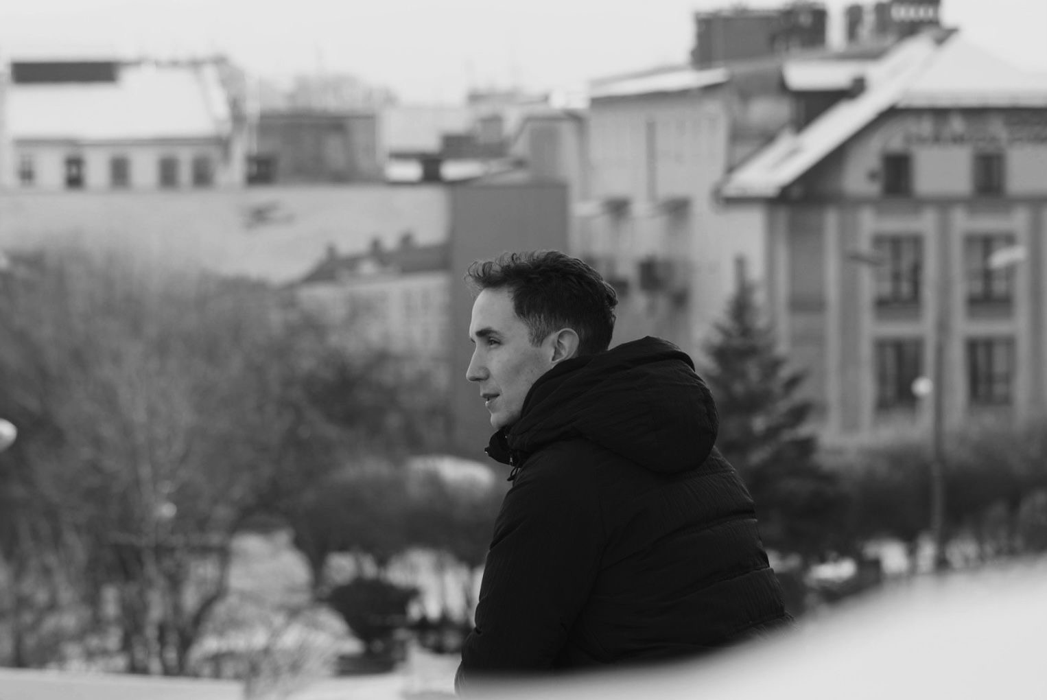 Czarno-białe zdjęcie profilowe mężczyzny w kurtce z kapturem, spoglądającego w dal na tle miejskiego krajobrazu z budynkami pokrytymi śniegiem.