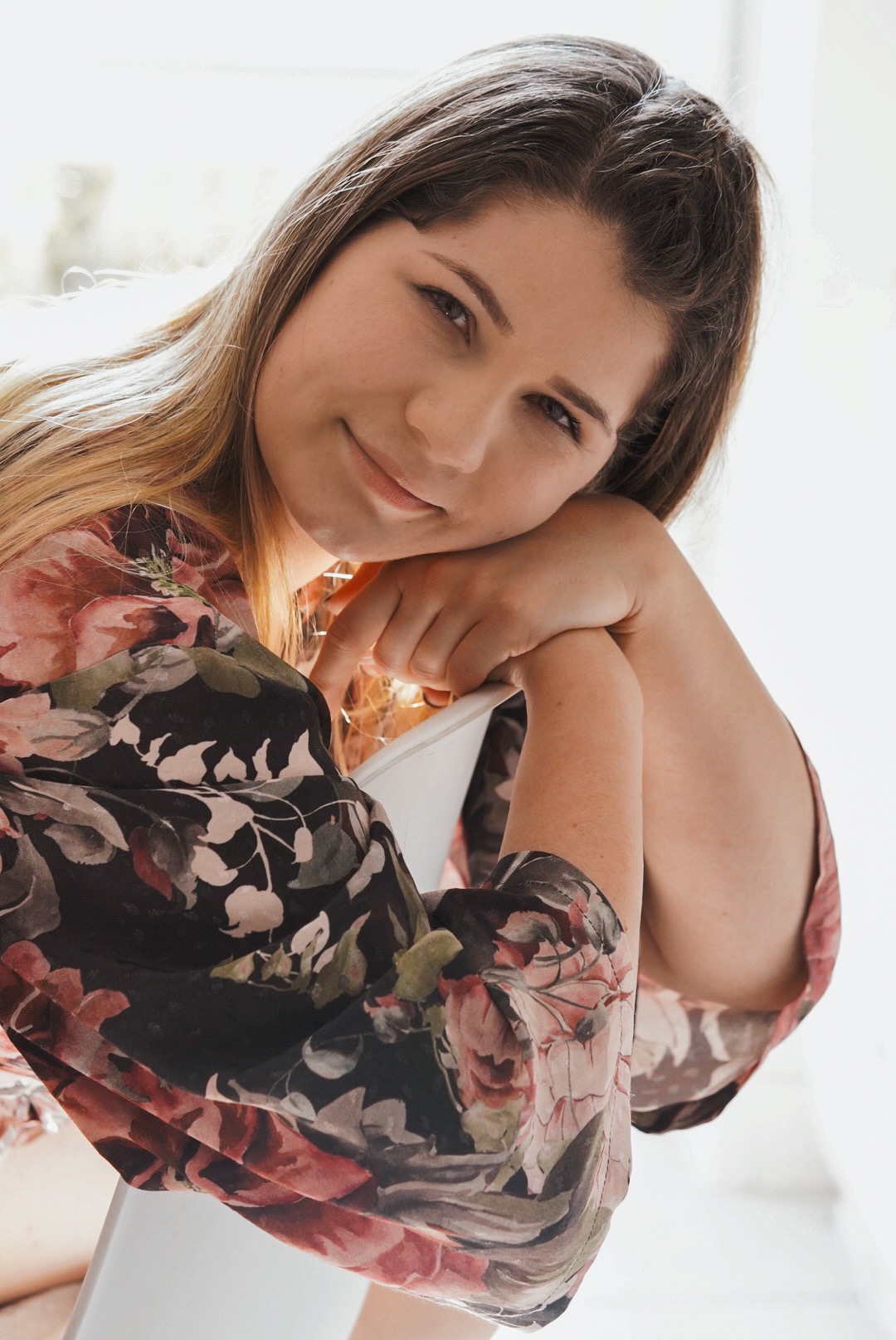 Portret kobiety w kwiecistej szacie, opierającej się na białym krześle, z delikatnym uśmiechem i spojrzeniem w obiektyw.