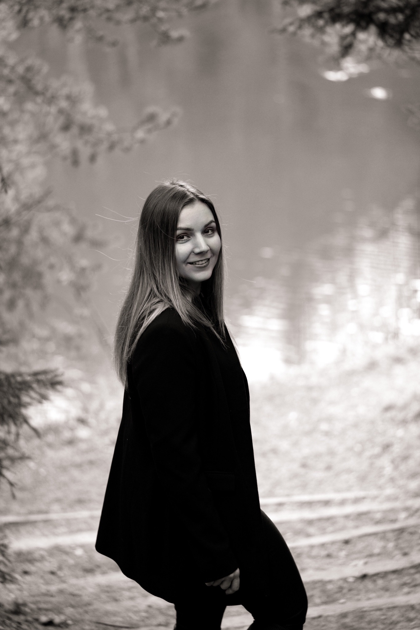 Portret uśmiechniętej kobiety w ciemnym płaszczu na tle jeziora i drzew w czerni i bieli.