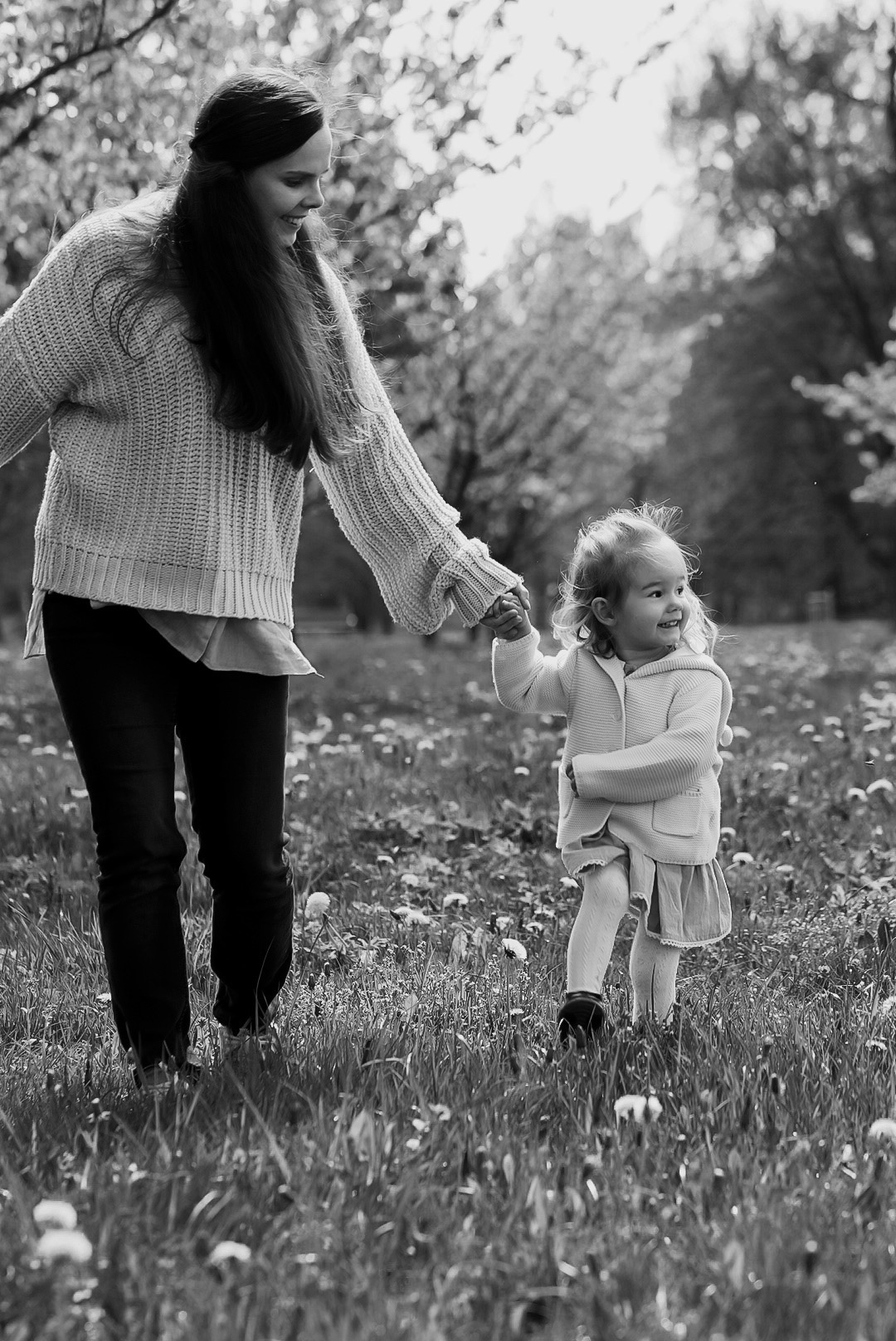 Czarno-białe zdjęcie matki trzymającej za rękę małą córeczkę na łące pełnej dmuchawców, obie uśmiechnięte, w tle drzewa.