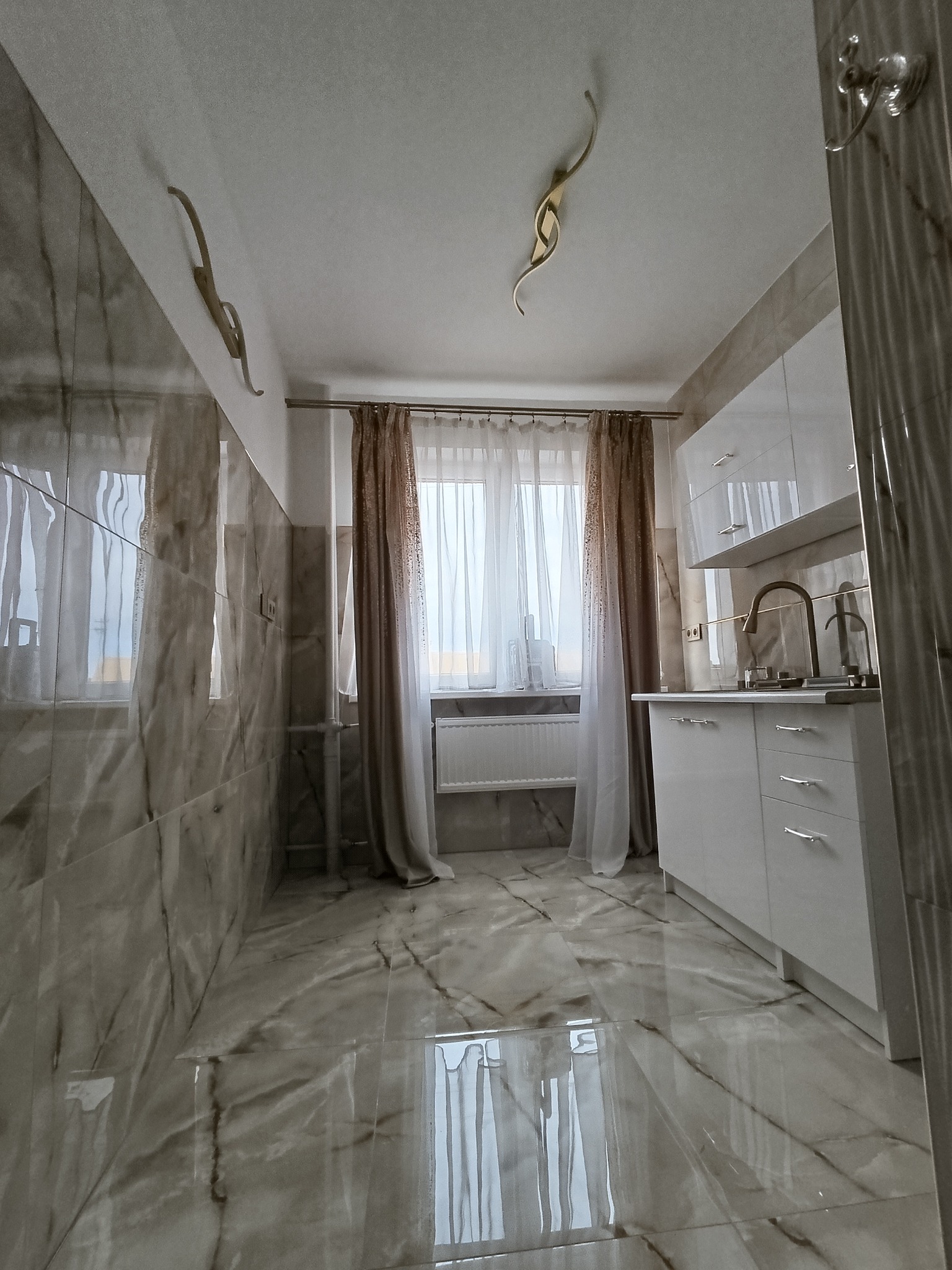 Wykończona kuchnia z błyszczącymi, marmurkowymi płytkami na ścianach i podłodze, białe szafki kuchenne z chromowanymi uchwytami, okno z białymi i brązowymi zasłonami, dekoracyjne, złote elementy...