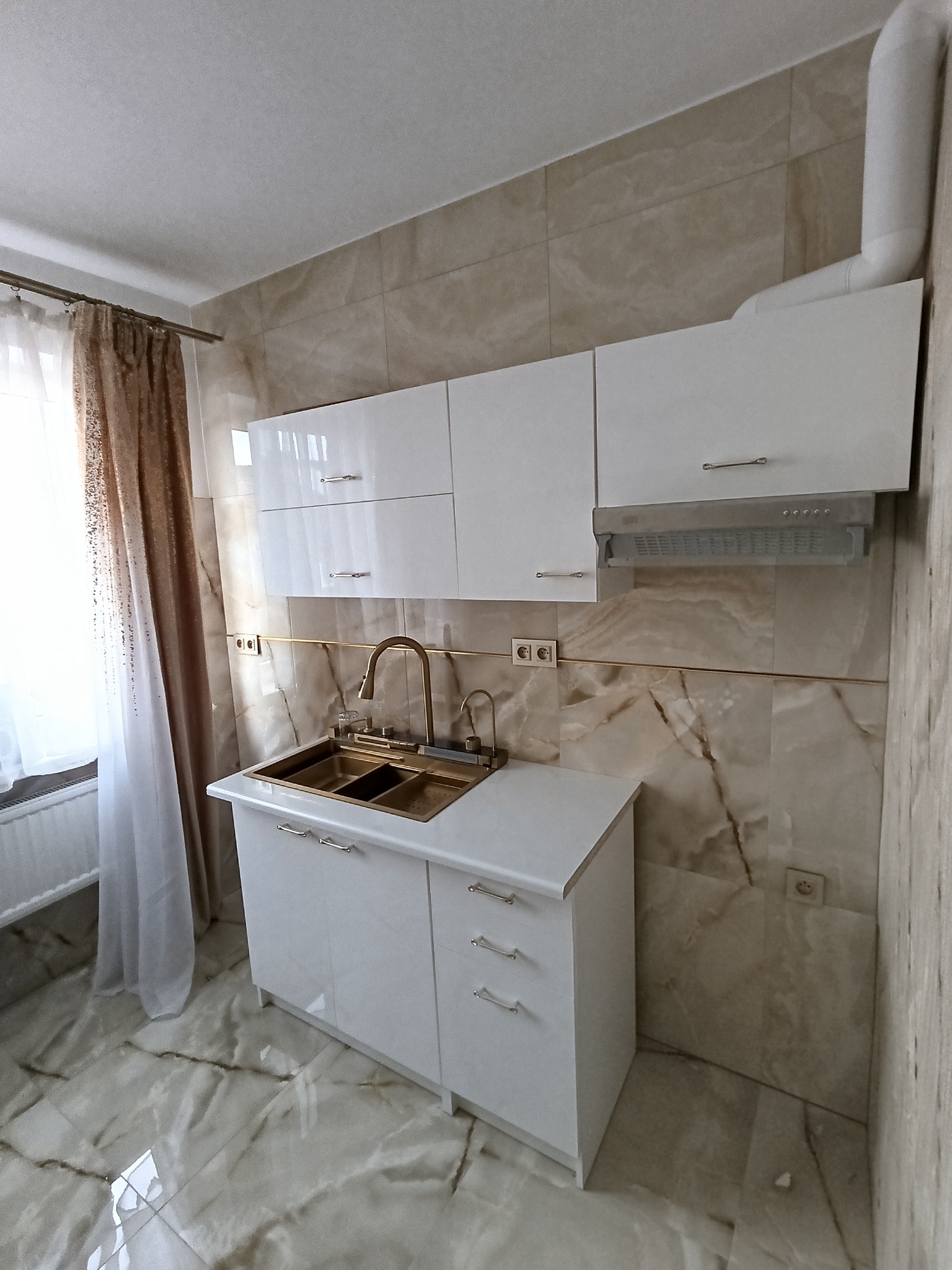 Nowoczesna kuchnia z białymi, lakierowanymi szafkami, złotą armaturą i marmurowymi płytkami na ścianie i podłodze, z widocznym oknem z zasłoną po lewej stronie.