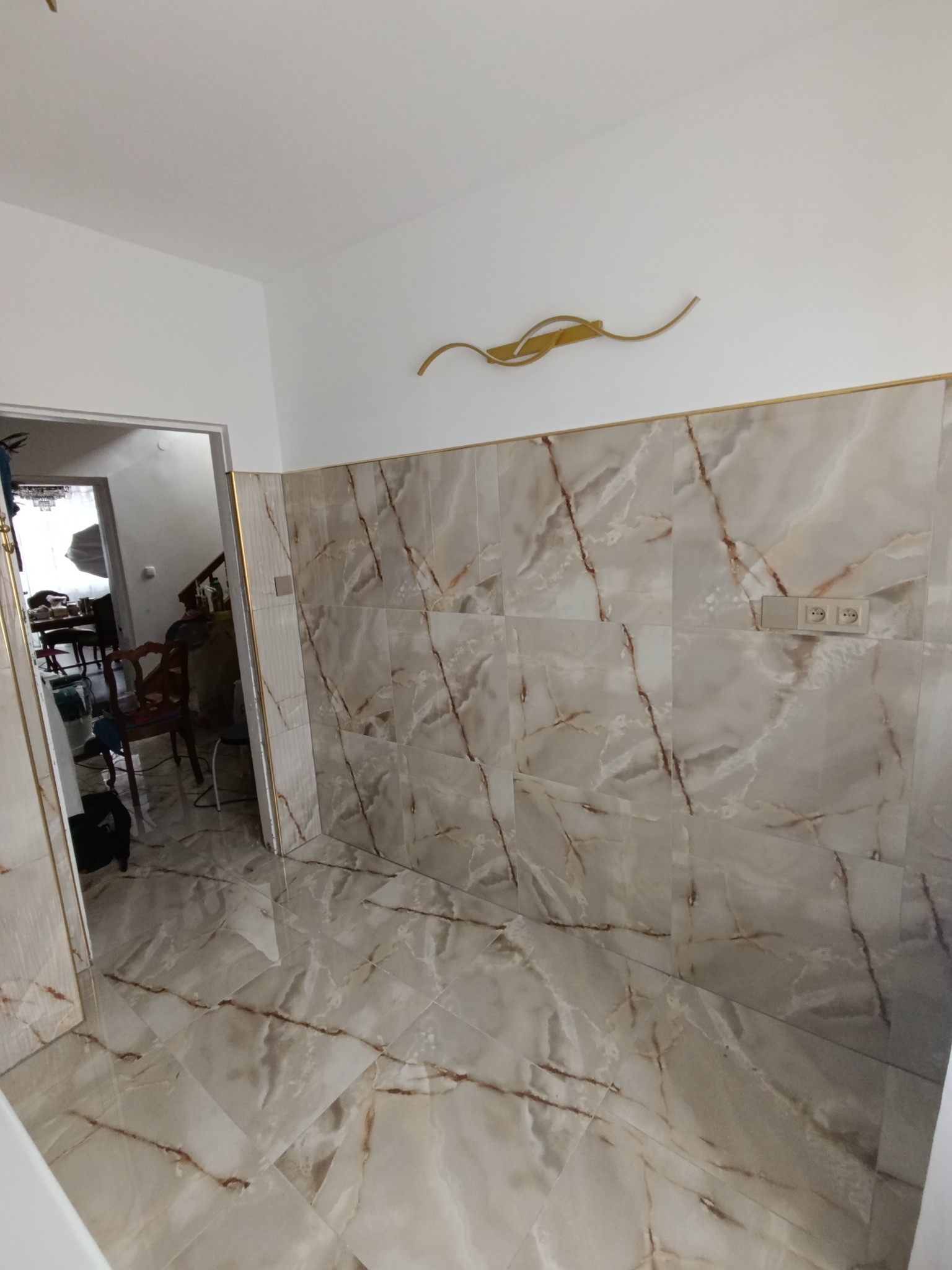 Wykończona łazienka z beżowymi płytkami imitującymi marmur na ścianach i podłodze, złote akcenty na krawędziach płytek i dekoracja ścienna, widoczne gniazdka elektryczne i otwarte drzwi do innego...