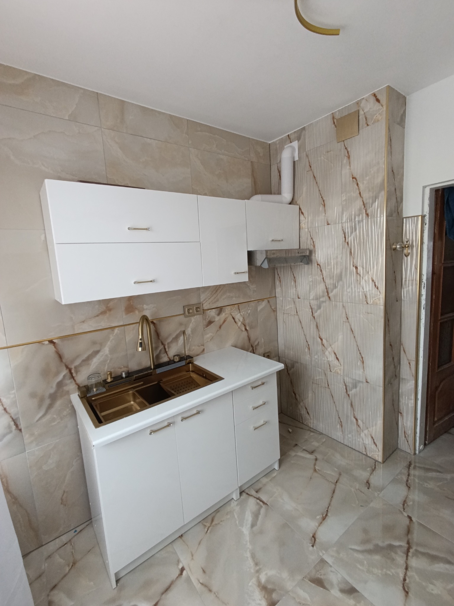 Wykończona kuchnia z marmurowymi płytkami, białymi szafkami z złotymi uchwytami i złotą armaturą, widoczny fragment wentylacji.