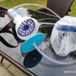 Prototypowy system filtracji powietrza z elementami HEPA, rurkami i maską ochronną, leżący na szklanym stole z wiklinową ramą, na tle zieleni ogrodu.